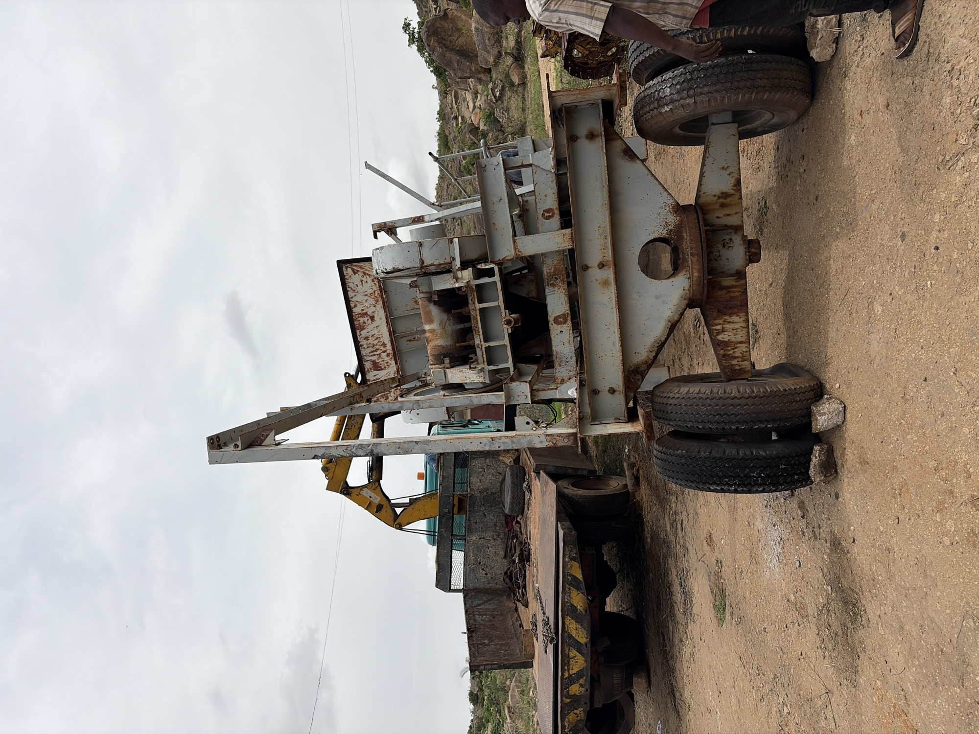 The image depicts a stationary, heavily rusted, industrial machine mounted on a flatbed trailer in what appears to be an outdoor setting near Gura Top, Nigeria. The machine, predominantly gray and brown with visible corrosion, features an intricate framework of metal beams and a complex inner structure. A yellow hydraulic arm is partially visible. The trailer, also rusted, has a black and yellow hazard-striped rear. The trailer's wheels are dual tires, some are flat, and appear damaged. An individual wearing a striped shirt and dark trousers stands partially in the top-right corner of the frame. The ground is dirt and gravel. In the background, there are rocky hills and a cloudy sky.