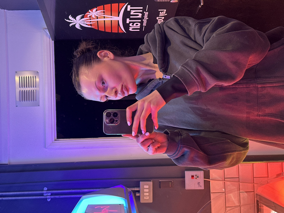 The image displays a person, likely female, taking a mirror selfie in what appears to be an indoor commercial setting, possibly a tanning salon or a similar establishment, given the visible signage. The photo is taken from a slightly high angle, looking down towards the person.
The subject is positioned in the right half of the frame, holding a dark-colored smartphone with multiple camera lenses in her hands, reflecting her face on its screen. She has dark hair, possibly pulled back, and is wearing a dark, full-zip hooded sweatshirt over a long-sleeved dark top. Her fingernails are painted a light pink or lavender color. Her expression is neutral.
The background is dimly lit, with prominent artificial lighting creating a moody atmosphere. To the left of the person's head, a black banner or sign is visible, featuring a graphic of a sunset with palm trees and the mirrored text "Tun Tan" above smaller, also mirrored text that appears to be "La Dolce Vita" (partially visible as "lɐ Do"). Above and to the far left of the person, on a white wall, there is a rectangular metal ventilation grate.
Below the person and towards the bottom-left of the frame, a bright blue and purple glowing object, possibly a neon light or part of a machine, casts a strong hue. A warm red or orange light emanates from the lower right side of the image, illuminating the person's right side and parts of the wall. On the wall to the person's right, several white and grey wall-mounted control panels or switches are visible, one with a small red light. Below these, the wall is covered with white rectangular tiles.
The overall impression is a casual, indoor scene, likely taken at night or in a windowless space, emphasizing the artificial colored lighting.