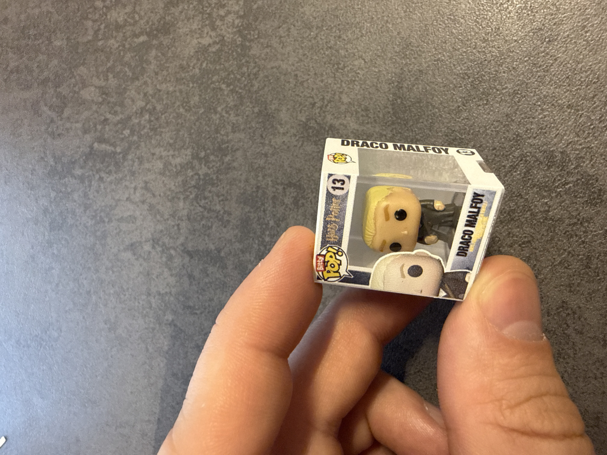 A hand is holding a miniature Draco Malfoy Funko Pop toy in its box. The box is white with black text and features a window showcasing the Draco Malfoy figure. The background is a gray textured surface. There is no indication of the location being in the city of Krasnoyarsk, Russia, or any specific location.