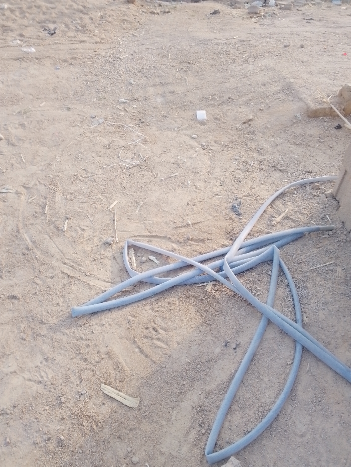 The image displays an outdoor ground scene, dominated by sandy and dusty terrain. The primary subject is a light blue, flexible hose or pipe, coiled and spread across the lower right and central portions of the frame. The hose appears to have a slightly textured or woven surface.
The ground is a muted beige-brown, indicative of dry earth, and shows minor irregularities and scattered small debris. Among the debris are small pieces of wood, visible towards the bottom left and near the hose. A small, white square object, possibly plastic or paper, lies on the ground towards the upper middle of the frame. Faint impressions or tracks are visible on the sandy surface.
In the background on the far right, a partial view of a light-colored, possibly concrete or stone structure is visible, suggesting a construction or undeveloped area. The overall lighting indicates daytime, with bright, natural illumination casting soft or no discernible shadows. The weather appears clear and dry. No individuals are present in the scene, and no specific activity is occurring. There is no visible text within the image. The setting is consistent with an outdoor, potentially utilitarian or undeveloped area in Panshekara, Nigeria.