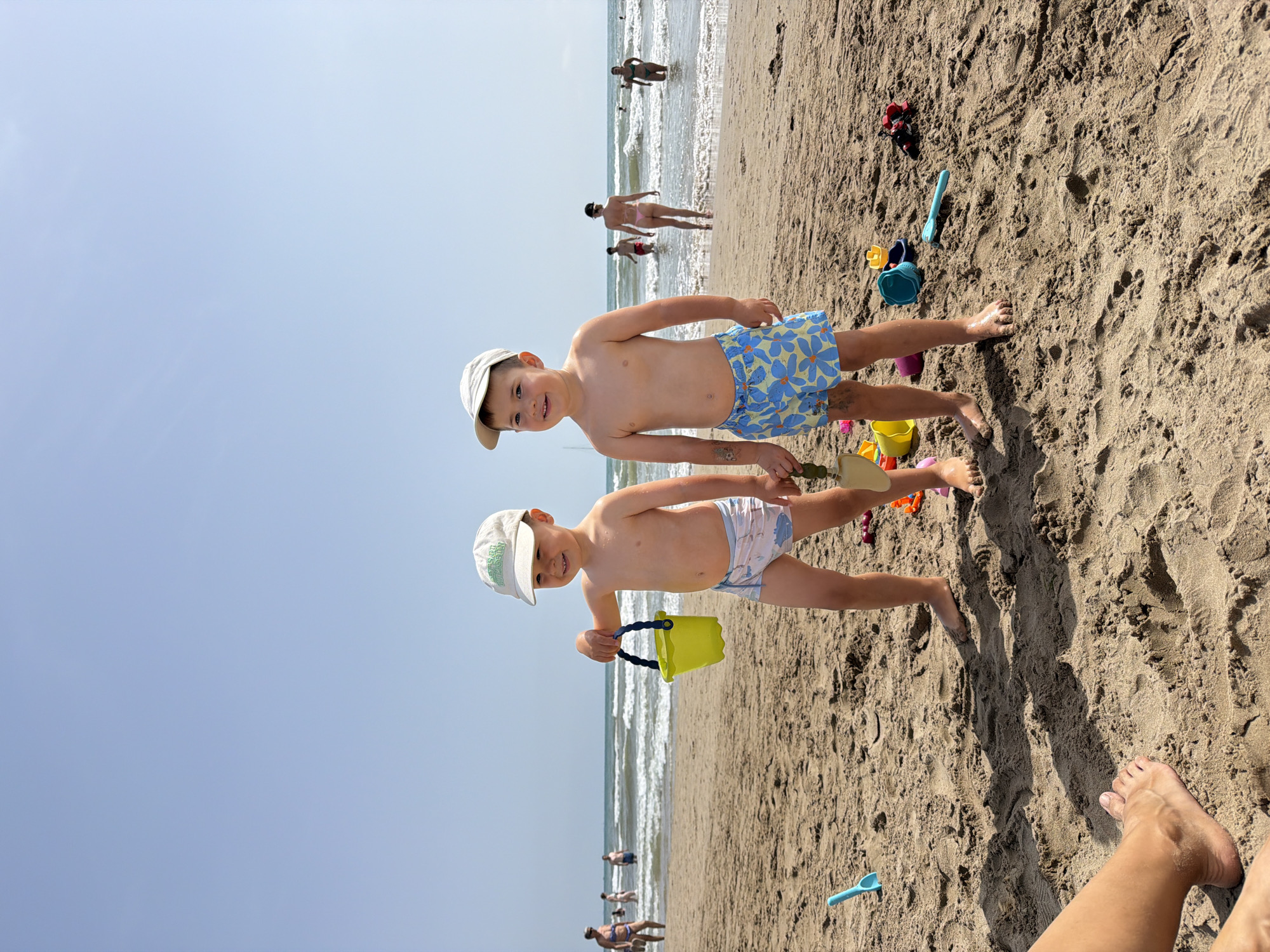 Two young boys wearing swim trunks and baseball caps stand on a sandy beach. The boy on the left, wearing a light-colored cap and patterned swim trunks, holds a yellow bucket. The boy on the right, with a similar cap and blue floral swim trunks, holds a toy shovel. In the background, the ocean has waves, and several other people are visible wading or swimming in the water. The sand is marked with shadows and indentations, suggesting the presence of sandcastles or other structures. Beach toys are scattered on the sand around the boys. The sky is clear and blue, indicating a sunny day.