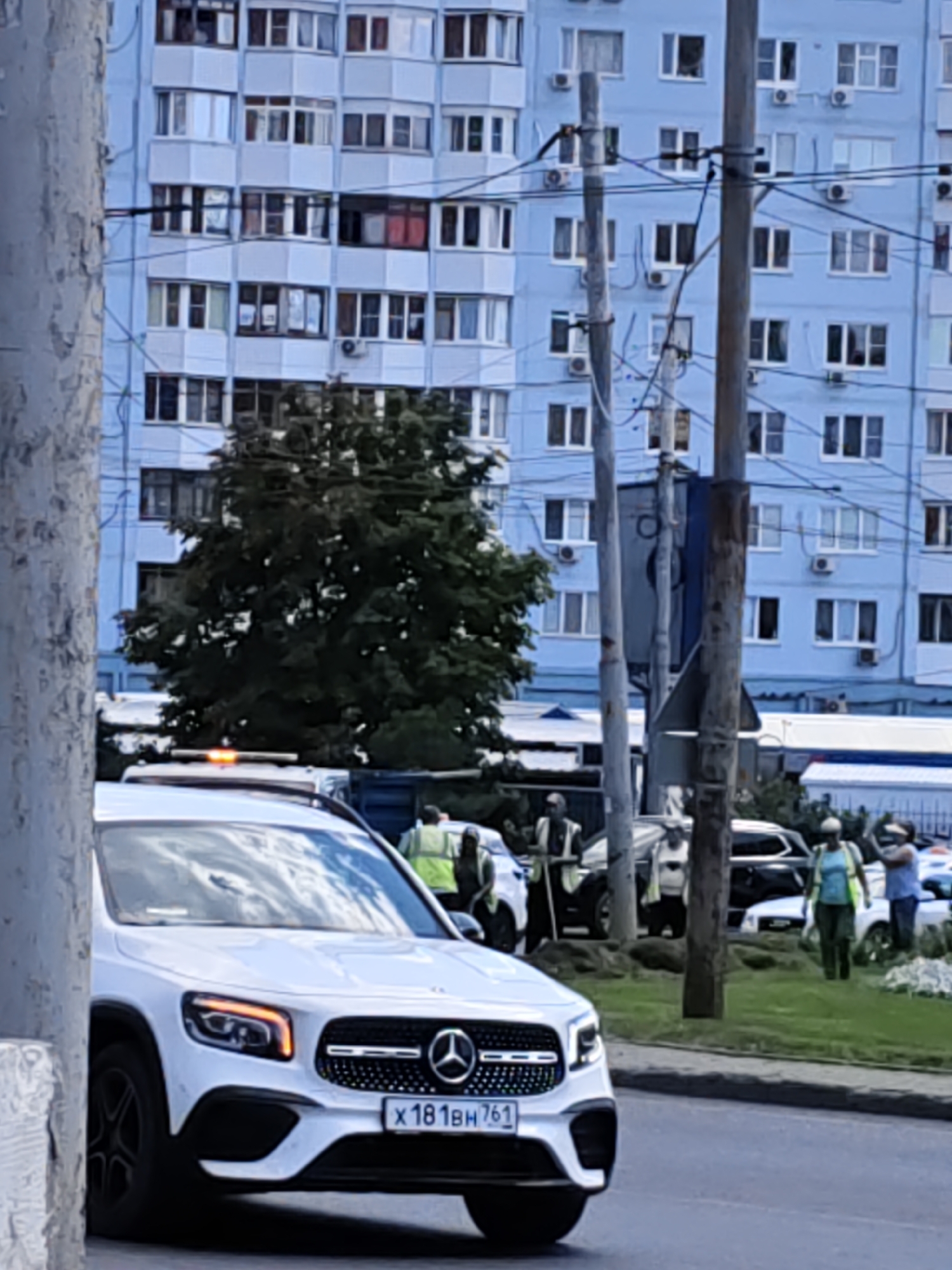 The image depicts a street scene in Rostov-on-Don, Russia. In the foreground, a white Mercedes-Benz car is visible. Behind it, several people are gathered, some wearing high-visibility vests, suggesting they might be road workers or emergency personnel. There is a large, light blue apartment building in the background, typical of urban architecture in the region. Trees and power lines are also present, contributing to the urban environment. The overall atmosphere appears to be that of an active street, possibly with some sort of work or event taking place. The weather appears to be overcast.