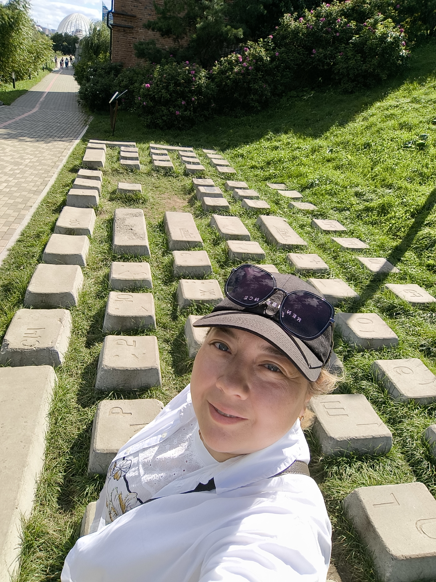 In Yekaterinburg, Russia, a woman wearing a white shirt, a dark baseball cap, and sunglasses is taking a selfie outdoors on a sunny day. She is positioned in the foreground, with her face partially visible. Behind her, a pathway paved with grey square tiles leads away into the distance. To the right of the path, a grassy slope is dotted with numerous large, rectangular concrete blocks arranged in rows. Many of these blocks have engraved symbols or letters on their surfaces, resembling oversized keyboard keys. The background features lush green trees, bushes with pink flowers, and a large, domed building. The overall environment suggests an outdoor public space, possibly a park or a monument.