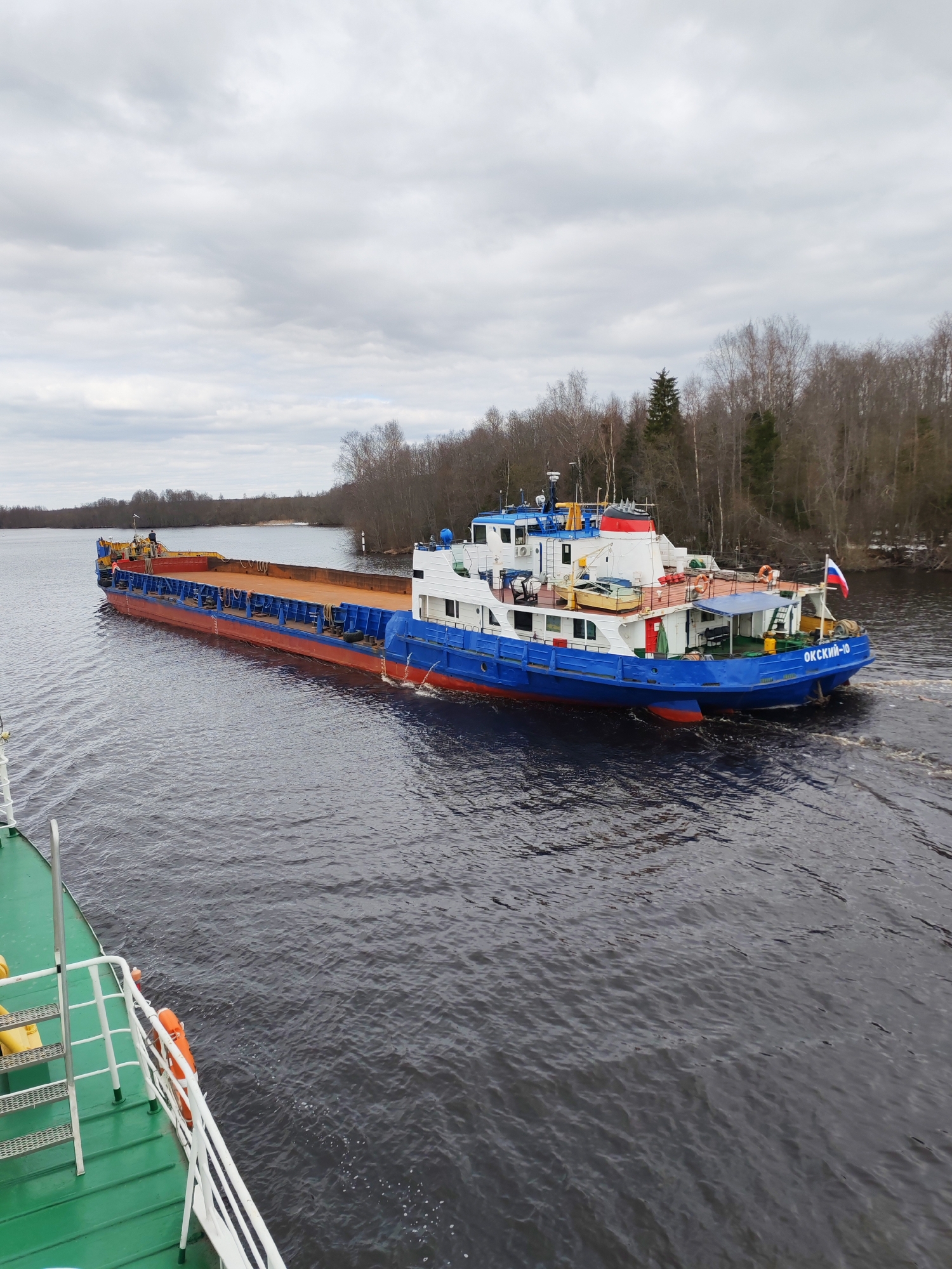 The image depicts a working vessel convoy navigating an inland waterway, likely a river or canal, in Uzhla, Russia. In the mid-ground, a blue and white tugboat or pushboat, identified by the name "ОКСКИЙ-10" (OKSKIY-10) on its stern, is propelling a long, empty deck barge. The barge has a red hull bottom, blue sides, and a flat, rust-colored deck. A single person is faintly visible near the bow of the barge. The tugboat flies a Russian flag from its stern.
The photograph is taken from the deck of another vessel, the green deck and white railings of which are visible in the lower left and bottom right corners. The water is dark and shows ripples and foam, indicating movement. The banks of the waterway on both sides are lined with a mix of bare deciduous trees and some evergreen trees, suggesting the season is late autumn, winter, or early spring when foliage is minimal. The sky is overcast with a uniform layer of grey clouds, providing soft, diffused light but indicating a dull day. There are no direct sunlight effects visible. The scene portrays a routine commercial transport operation on a calm, natural waterway.
