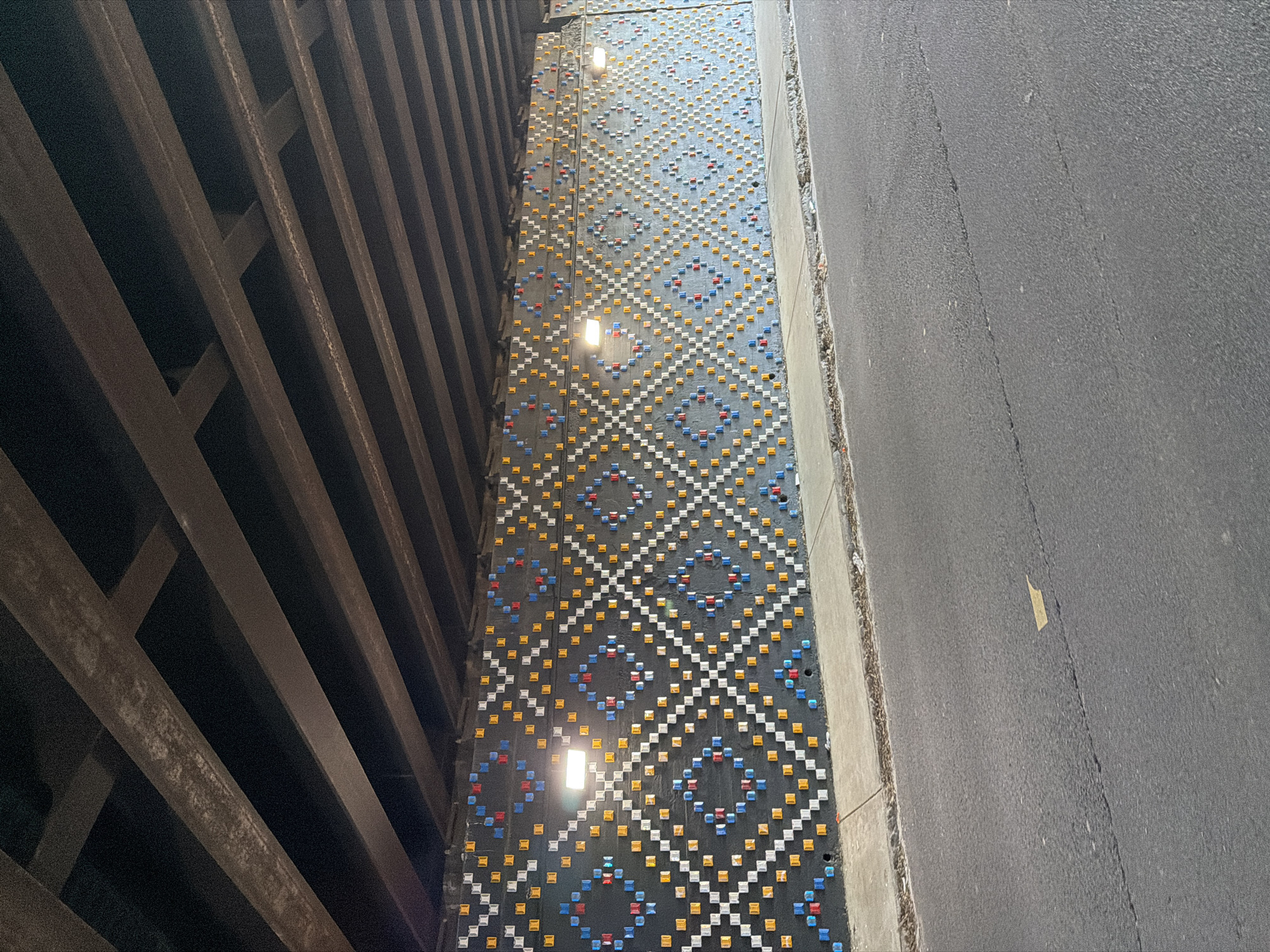 The photograph, taken in Atlanta, United States of America, presents a view looking down into a narrow passageway beneath what appears to be a bridge or overpass. On the left is a steel structure composed of intersecting beams and on the right is a gray asphalt surface, likely a road. Between the steel structure and the road is a narrow wall adorned with a geometric pattern of colored squares, primarily white, yellow, blue, and red. Three illuminated lights are positioned along this wall, their placement suggesting a pattern or design element. The overall composition is a study of urban infrastructure and the intersection of design, structure, and transportation.