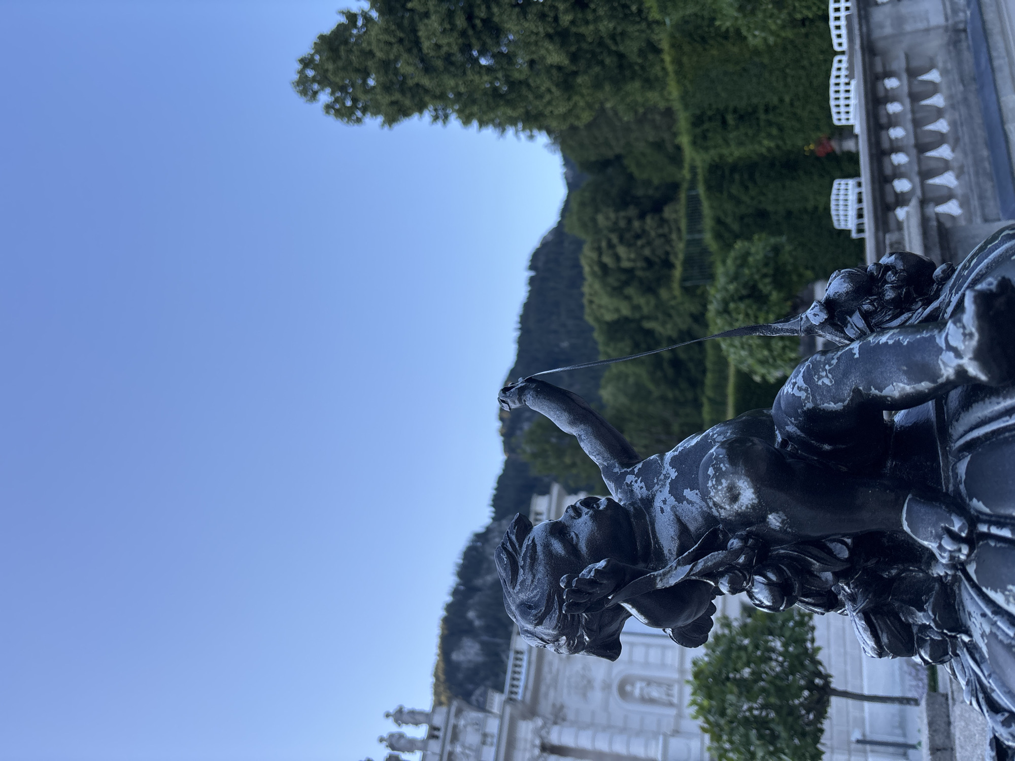 The image depicts a dark, weathered statue of a cherub in the foreground, appearing to be a fountain element. The cherub is positioned on a decorative base, holding an outstretched arm with a thin rod or stream of water. Behind the statue, a grand building with classical architecture, possibly a palace or monastery, is visible to the left. The background features lush green trees and hedges, with a mountainous landscape and a clear blue sky in the distance. The scene is likely an outdoor garden or courtyard setting, possibly at Linderhof Palace in Ettal, Germany, given the architectural style and the surrounding landscape. The lighting suggests it is either early morning or late afternoon.