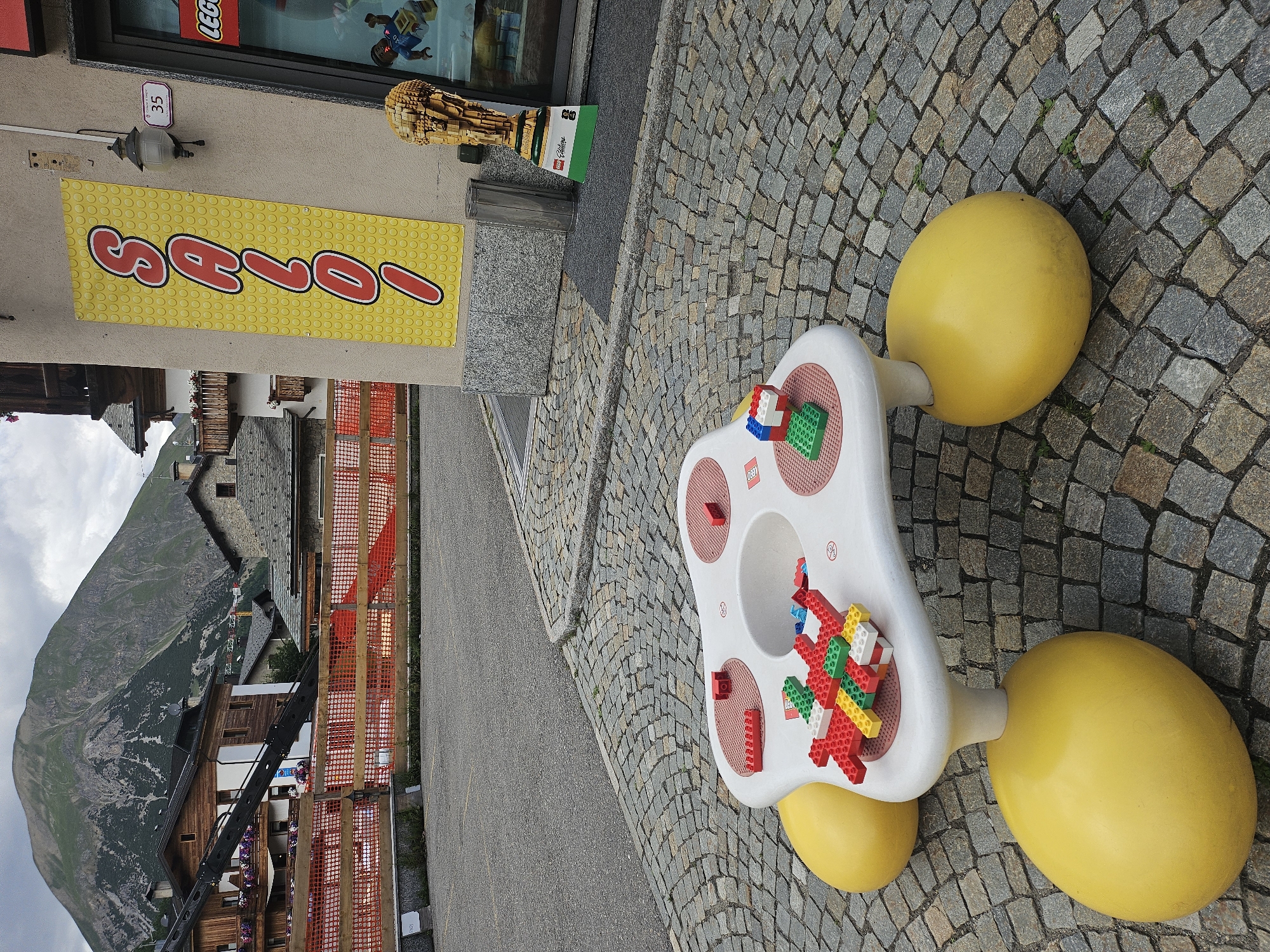 A bright yellow banner with red lettering, spelling out "SAVOIA", is displayed on a light-colored wall. The banner is adorned with a pattern of yellow circles. Above the banner, a small sign with the number "35" is visible, next to a decorative lamp. To the right of the banner, a tall, wooden sculpture shaped like a paintbrush, constructed from Lego bricks, stands prominently.
On a cobblestone pathway, a white Lego-themed activity table is set up. The table features red circular indentations and has Lego bricks in various colors, including red, green, and white, assembled on its surface. Two large, yellow, egg-shaped objects are attached to the base of the table, resembling wheels. The surrounding area is paved with gray cobblestones.
In the background, a picturesque mountainous landscape unfolds under a cloudy sky. Wooden buildings with dark roofs are nestled among the mountains. Orange construction netting is visible along a pathway. The scene appears to be outdoors in a village setting. The overall impression is one of a playful and welcoming environment, likely a Lego store or a public space featuring Lego-themed attractions.