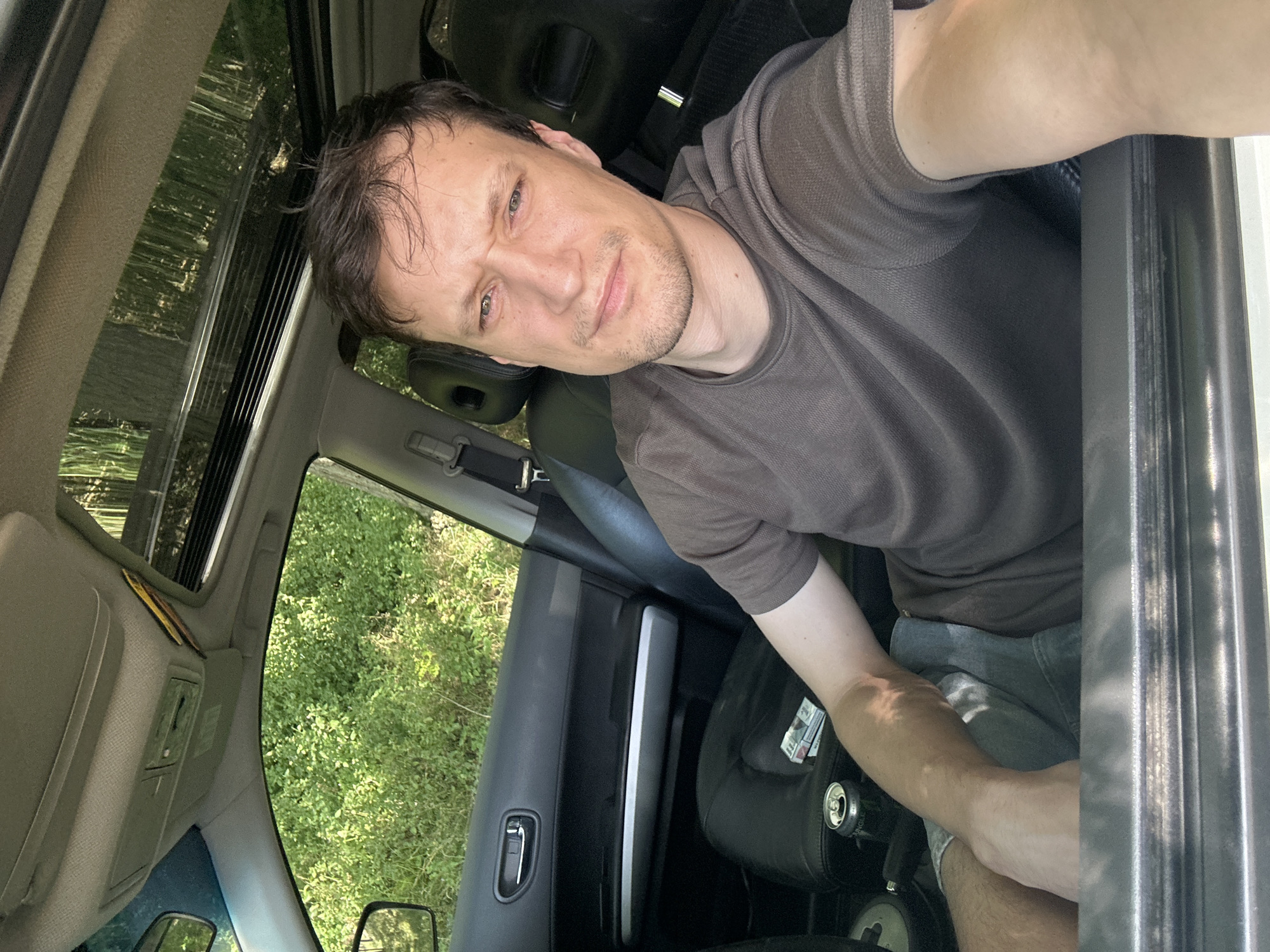 A man is sitting in the driver's seat of a car. The car's sunroof is open, and sunlight is filtering through the trees outside. The man is wearing a brown t-shirt and denim shorts. He has short brown hair and stubble on his face. There is a pack of cigarettes and a can of soda on the passenger seat. The car appears to be parked on a dirt road in a wooded area. The location is Makiv, Ukraine.