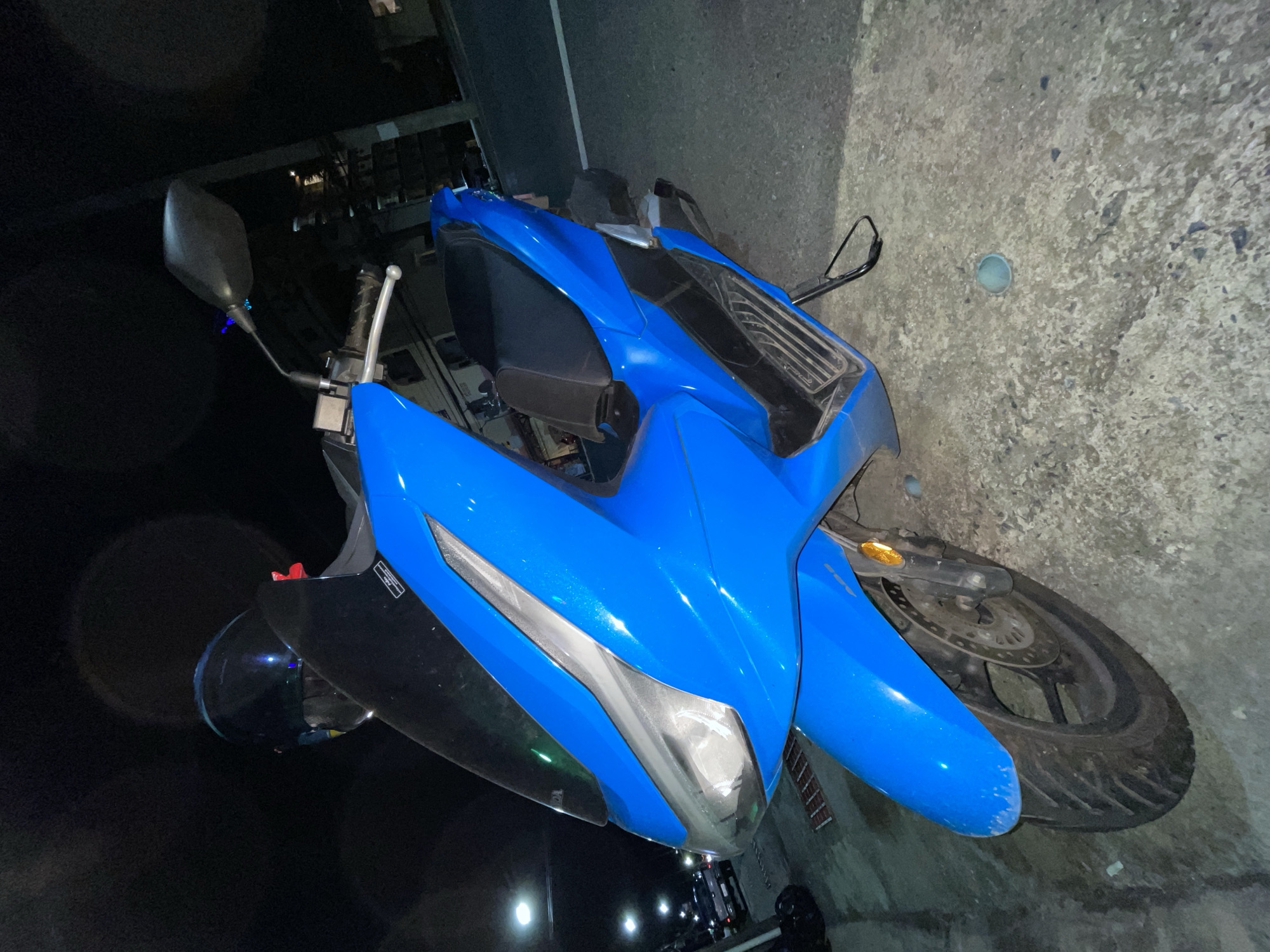 A blue scooter is parked on a concrete surface at night. The scooter has a black seat and a dark tinted windshield. A black helmet is placed on the handlebars. The background shows streetlights and buildings, suggesting an urban environment. The scene appears to be in the city of Chalong, Thailand.