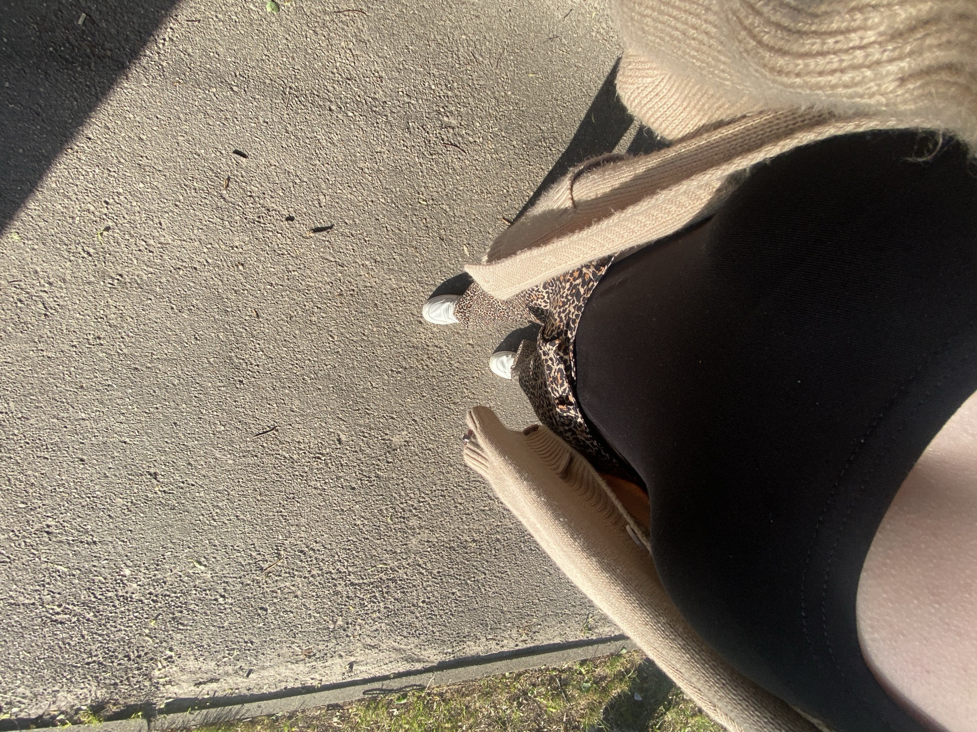 An overhead shot captures a person standing on an asphalt surface, looking down at their feet and lower body. The subject is partially visible, wearing a beige, chunky knit cardigan with a ribbed texture. Underneath the cardigan, a black garment, possibly leggings or a skirt, is worn over what appears to be a leopard print skirt or dress. White sneakers are visible on the person's feet.
The setting is outdoors, likely a paved path or sidewalk, with a concrete curb and a narrow strip of green grass visible at the very bottom edge of the frame. Strong sunlight casts sharp, dark shadows across the asphalt, suggesting a clear and bright day. The shadows indicate the sun is at an angle, possibly mid-morning or late afternoon. No specific activity beyond standing or walking is discernible. There is no visible text in the image.