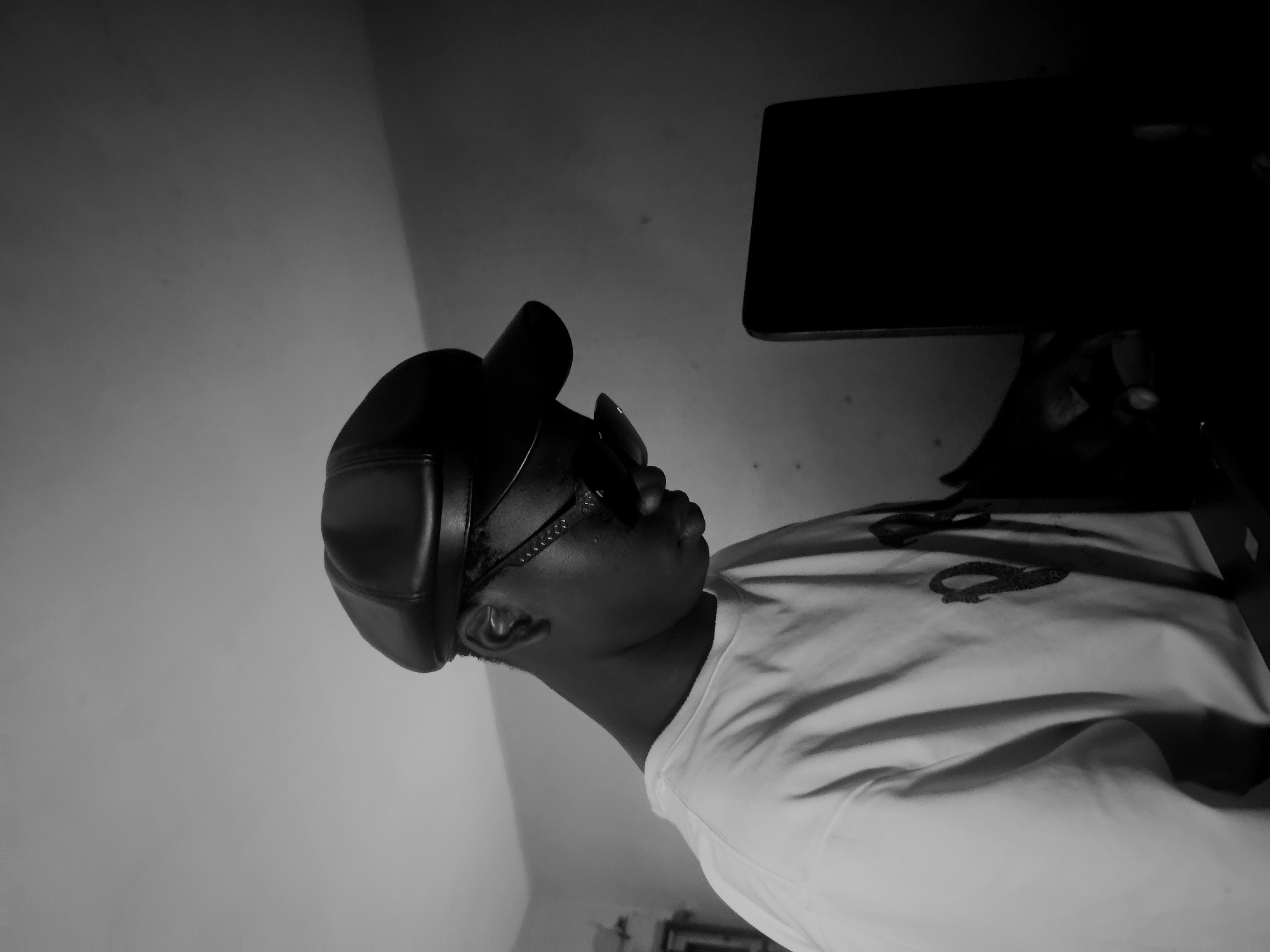 A man in a baseball cap and sunglasses stands in front of a table in Nkwelle, Nigeria. The image is in black and white. The man is wearing a white t-shirt.