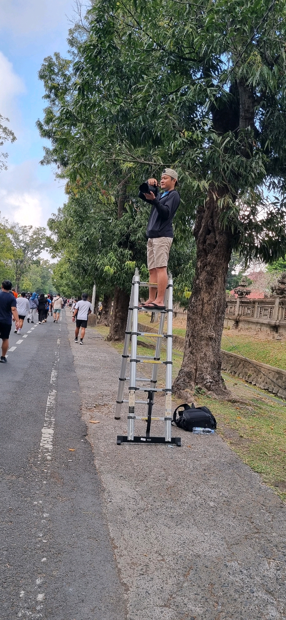 A man, wearing a baseball cap, dark long-sleeved shirt, and khaki shorts, stands on a silver, multi-section ladder and holds a black camera to his eye, appearing to take a photograph. The ladder is positioned on the edge of a paved walkway adjacent to a road. A black backpack and a clear plastic water bottle are on the ground next to the ladder. The walkway runs alongside a tree-lined street, with a line of people walking on the road in the background. A wall with decorative carvings is visible beyond the trees. The sky is partly cloudy. This scene is likely within the city of Denpasar, Indonesia, given the architectural style of the wall.
