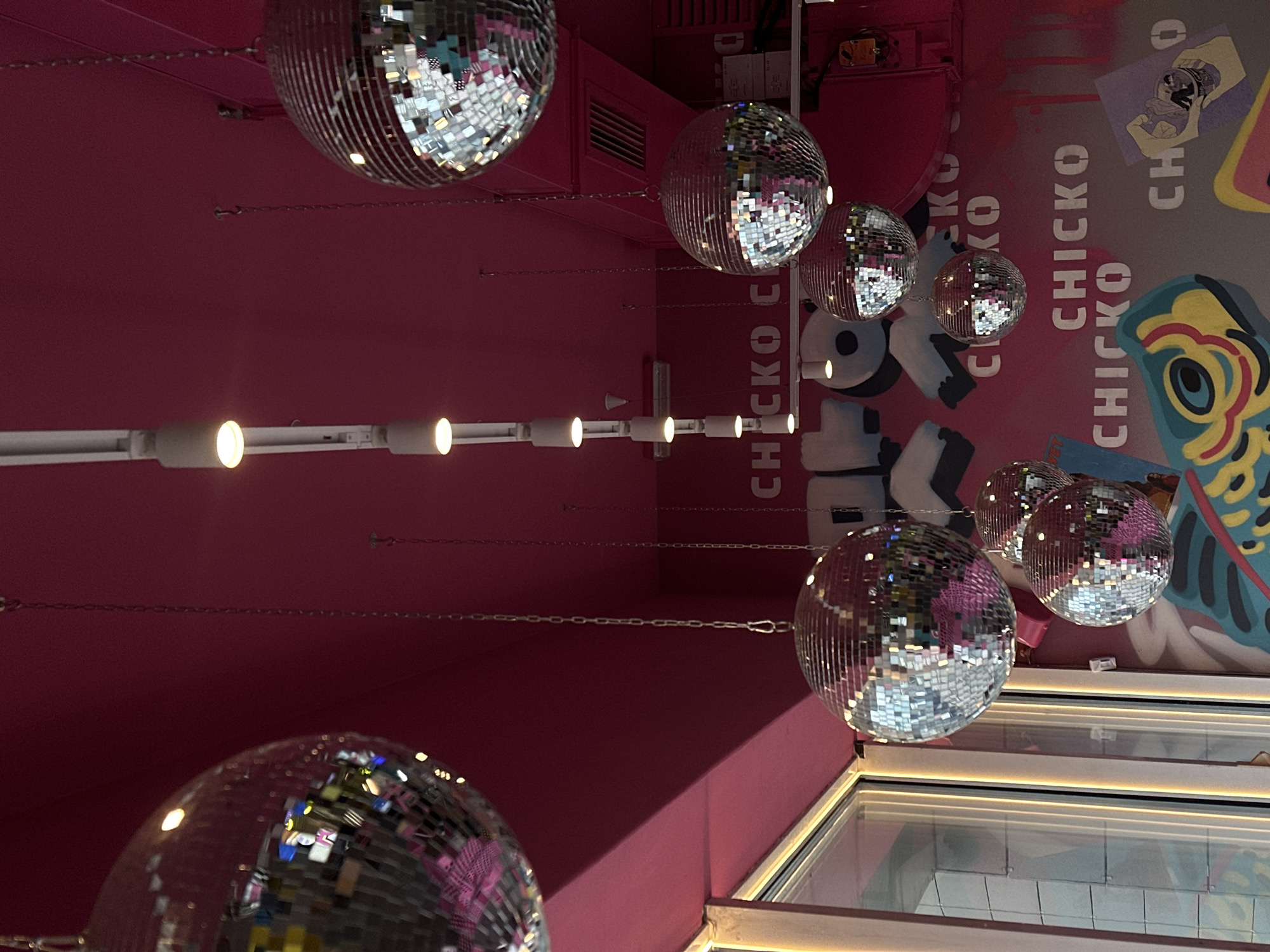 The image displays a brightly lit interior space, characterized by a vibrant pink and silver aesthetic, indicative of a modern commercial establishment in Perm, Russia. The perspective is slightly tilted, showcasing a solid pink wall on the left that meets a ceiling or an adjacent wall adorned with a mural on the right.
Numerous silver disco balls of various sizes are suspended from the ceiling, reflecting the artificial lights and surrounding colors. These disco balls are attached by thin metallic chains. Along the ceiling, a white track lighting system runs horizontally, equipped with several warm-toned spotlights that illuminate the space.
On the right side of the image, a colorful mural is prominently featured. The mural includes the word "CHICKO" stenciled repeatedly in white capital letters, sometimes partially obscured or overlapping. Other graphic elements within the mural include stylized, bold illustrations, such as a large, colorful design resembling a fish or character head in hues of blue, yellow, and red, along with abstract shapes and patterns. Below the mural, parts of what appear to be windows or display cases are visible, reflecting the interior lighting and contributing to the bright atmosphere.
The scene captures the unique decor of the venue, suggesting a lively and playful atmosphere. No specific activity is taking place, and no people are visible. The artificial lighting makes the time of day indistinguishable.