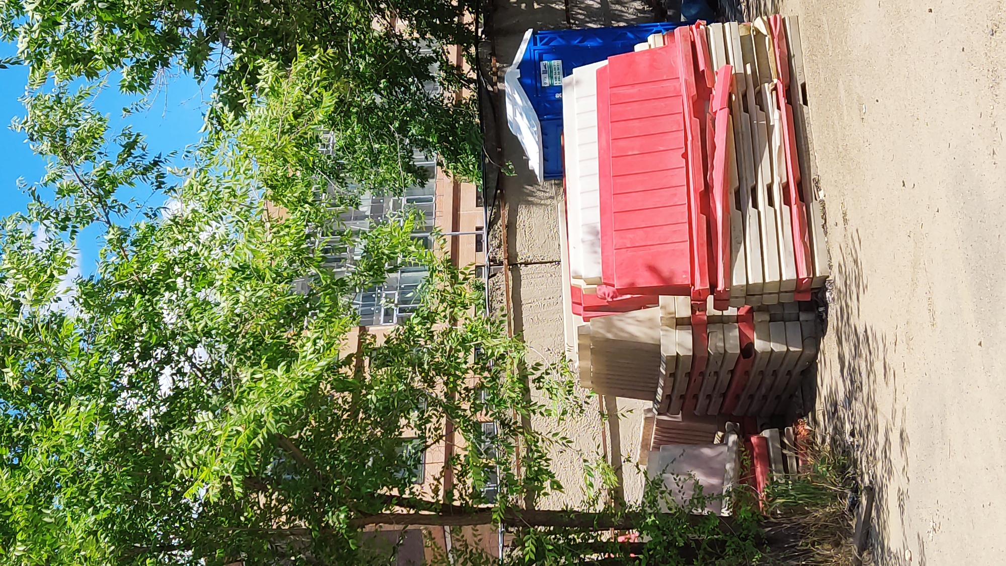 The image depicts an outdoor scene in Krasnodar, Russia, on a clear day with a bright blue sky visible through lush green tree foliage. In the foreground, stacks of red and white plastic barriers are arranged on a dirt surface. To the right, a blue portable toilet is situated behind the barriers. In the background, a multi-story building with numerous windows is visible, partially obscured by trees and a textured wall. The overall impression is of a construction or temporary event site.