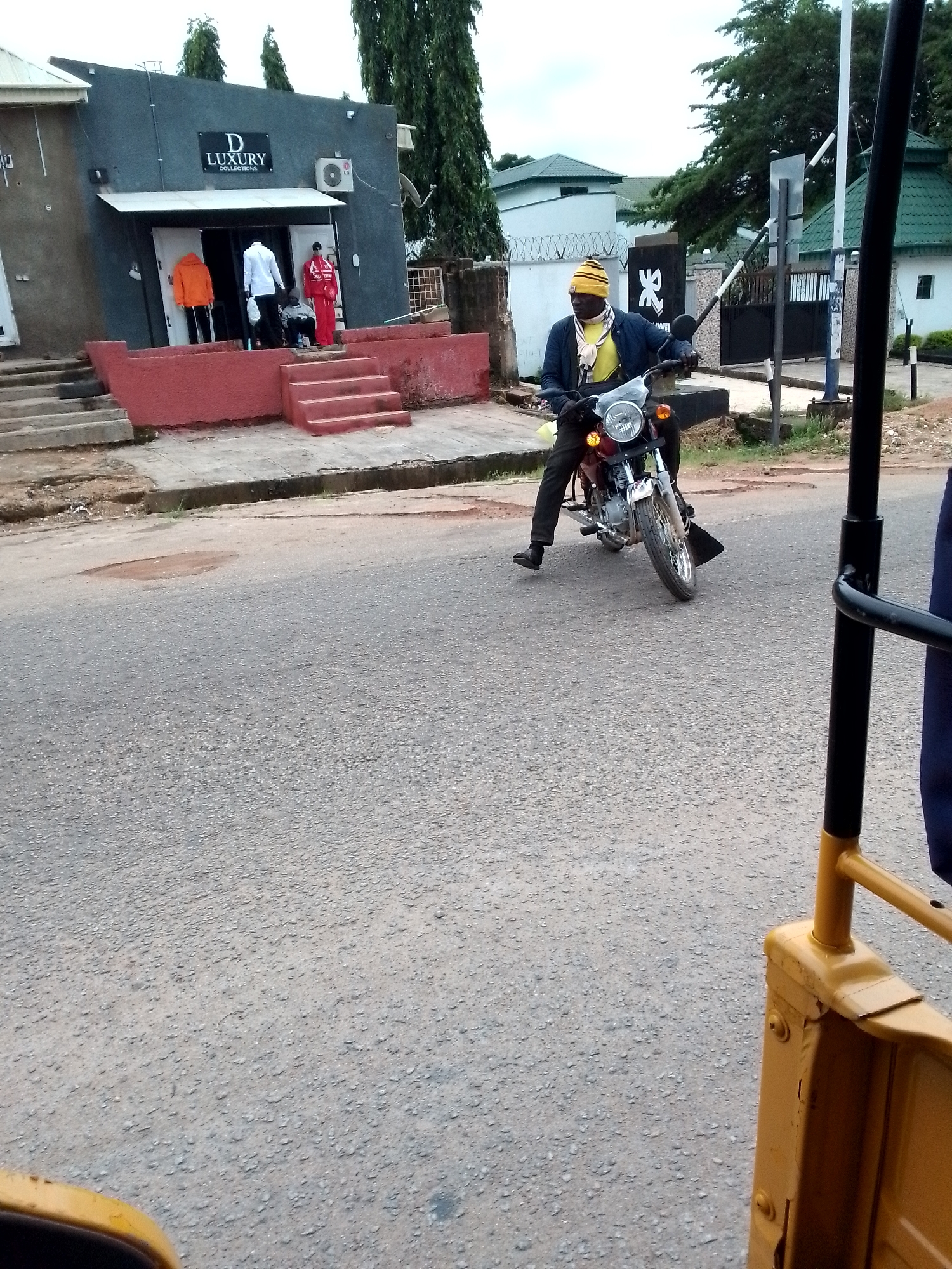 A man wearing a yellow and black striped beanie, a yellow t-shirt, a blue jacket, and dark pants is riding a motorcycle on a paved road. He is positioned in the center of the frame, facing the viewer's left. The motorcycle is red and black with a silver headlight. In the background, to the left, is a building with a sign that reads "D LUXURY COLLECTIONS." There are mannequins displayed in front of the building. To the right of the building, there are trees and a white fence with a gate. The overall scene appears to be in an urban environment, possibly a street in Jos, Nigeria, given the presence of a motorcycle commonly used as a taxi in that region. The lighting suggests it is daytime, and the sky is overcast. The foreground shows a yellow structure, possibly part of a vehicle or a barrier, suggesting the photo was taken from another moving vehicle.