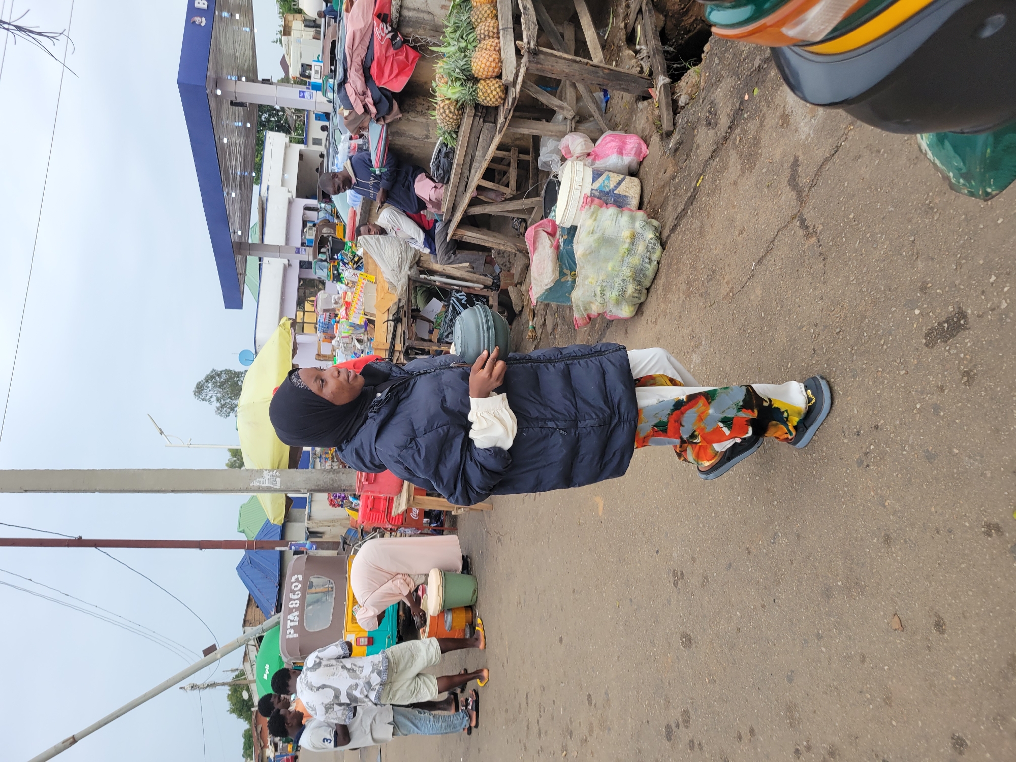 The image depicts a street scene in Jos, Nigeria. A woman wearing a black hijab, blue puffer jacket, white blouse, and patterned pants stands on a paved street, holding a green object. Behind her, a rickety wooden stand displays pineapples, clothing, and packaged goods. A gas station is visible in the background. Several people are present; some are near auto rickshaws (tuk-tuks), while others are tending to buckets. The weather appears overcast.