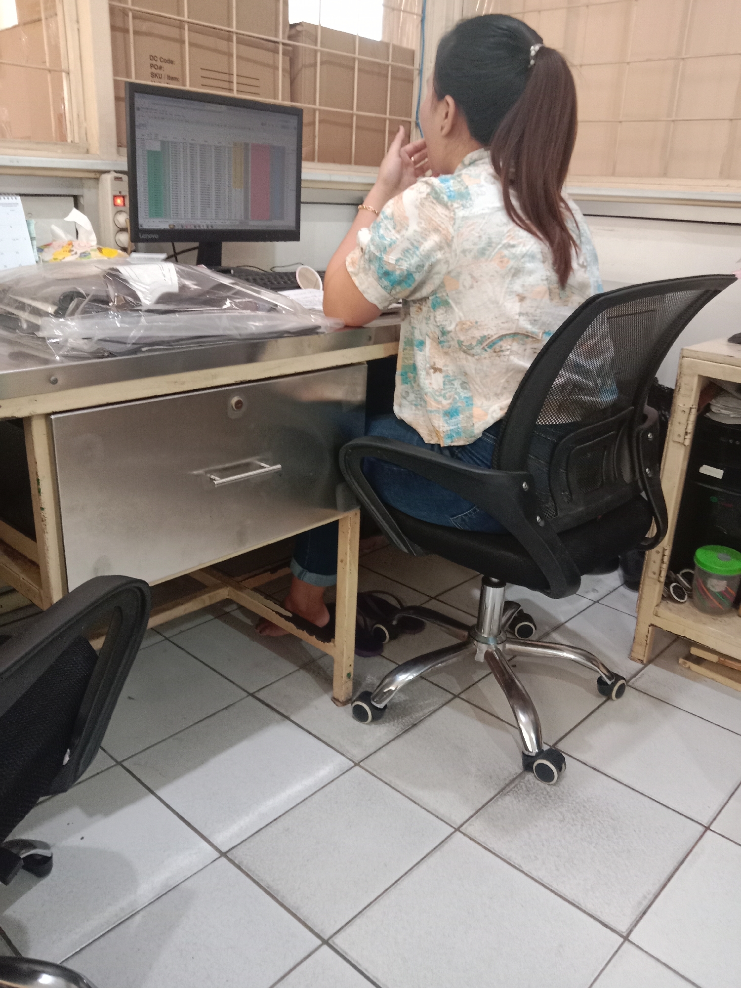 In an indoor setting, likely an office, a woman with her back to the camera is seated at a desk. She has dark hair pulled into a high ponytail and is wearing a light-colored, short-sleeved shirt with a patterned design and blue jeans. Her right hand is near her face, and she appears to be looking at a computer monitor on the desk. The monitor displays a spreadsheet with colored columns. To the left of the monitor is a calendar, a box of tissues, and some items covered in clear plastic sheeting. The desk itself is metallic with a prominent drawer. An office chair with a black mesh back and seat is positioned in front of the desk. The floor is tiled. In the background, there are shelves with boxes, and to the right, a cabinet with some items on it, including a green plastic container. This scene is consistent with an office environment, possibly within the city of Semarang, Indonesia, given the context of the request.