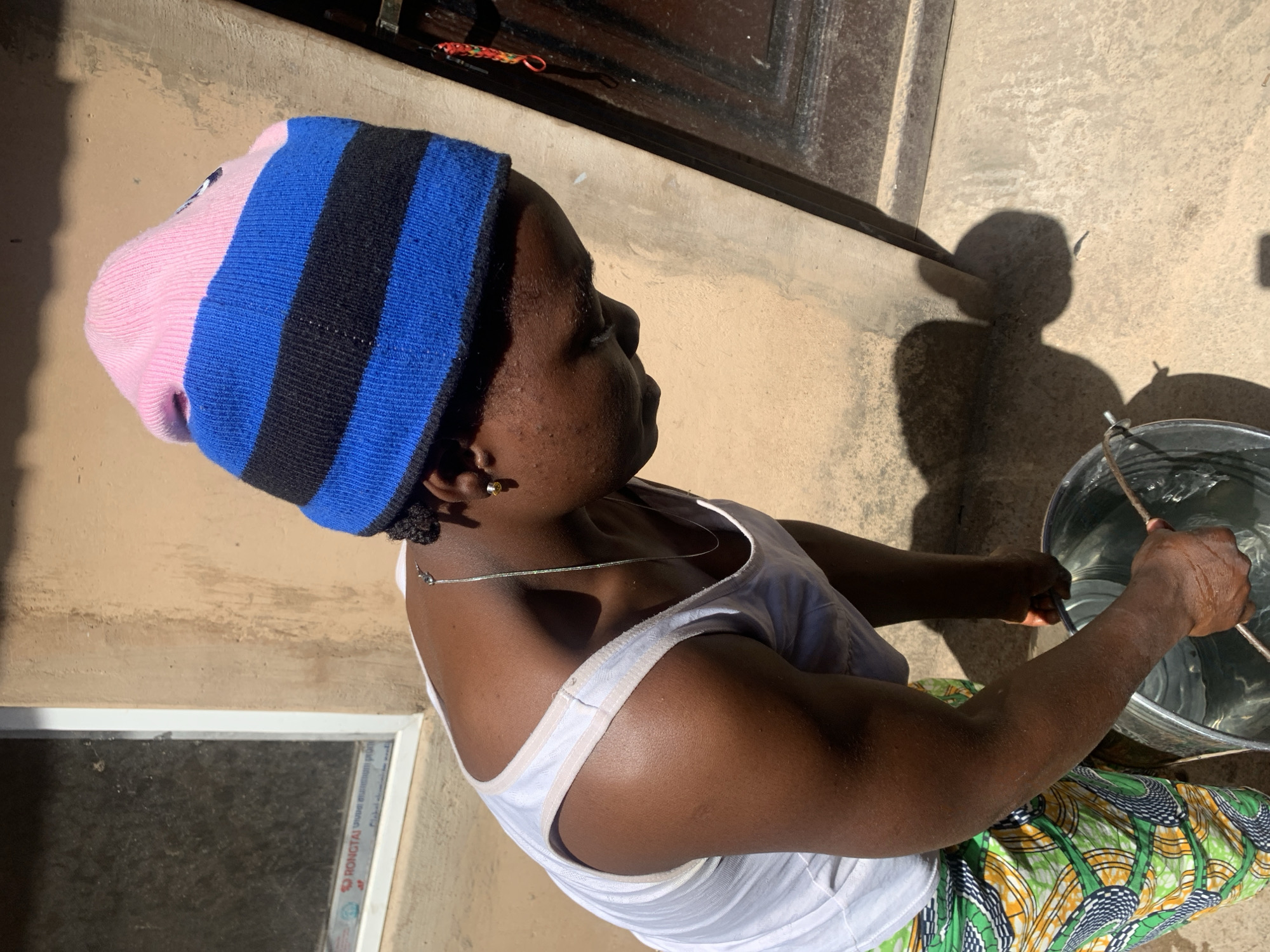 A medium-shot, portrait-oriented photograph depicts a person engaged in an outdoor activity under bright sunlight in Jos, Nigeria. The individual, with dark skin, is seen from the side, looking towards the right, wearing a vibrant knitted beanie with pink, blue, and black stripes. They wear a simple white sleeveless tank top and a patterned wrap skirt or cloth featuring green, yellow, and black designs. A thin silver chain necklace adorns their neck, and a small yellow stud earring is visible.
The person is actively using both hands to manipulate a stick or rod within a large metallic bucket filled with water, suggesting a task such as fetching, stirring, or washing. Their hands appear wet from the activity.
The setting is outdoors against a light tan-colored wall that shows some weathering. In the upper background, part of a dark window or door frame is visible. On the lower section of this frame, a white sticker with faint text is present, where "RONGTAI" and phrases like "Good material" can be partially discerned. Strong, direct sunlight casts a prominent and well-defined shadow of the person and the bucket onto the wall and the adjacent paved or concrete ground, indicating a clear and bright day, likely mid-day or afternoon. The overall scene captures a moment of daily life.