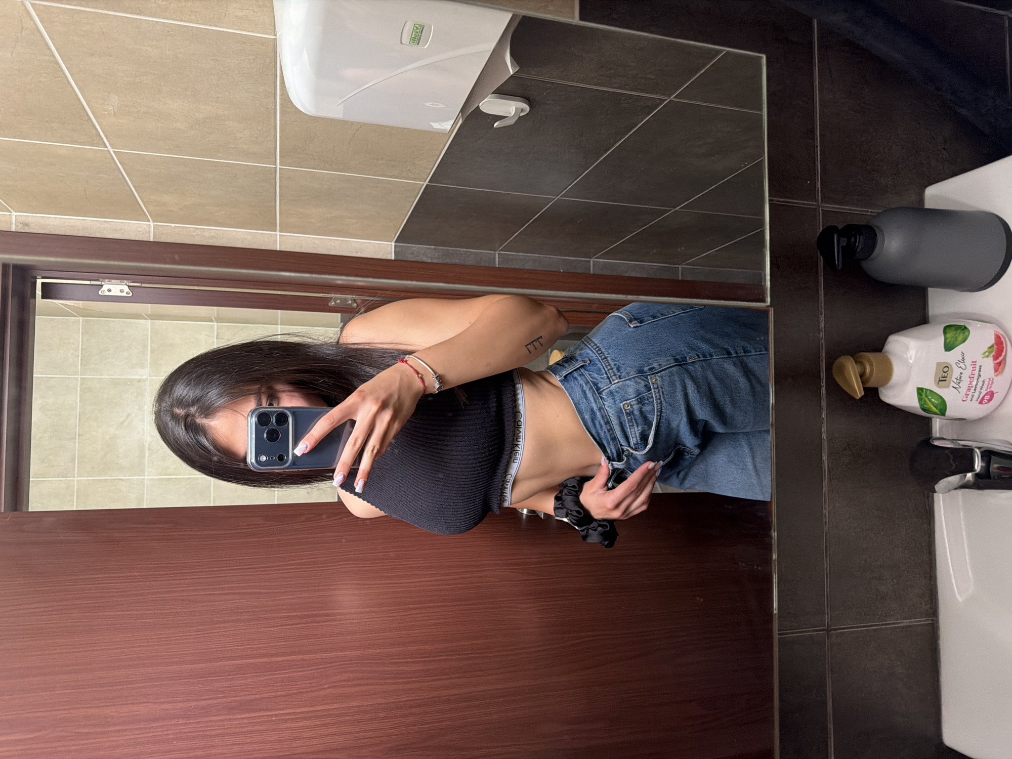 A young woman is captured taking a mirror selfie in what appears to be a bathroom, with the image rotated 90 degrees clockwise. She has long dark hair and her face is largely obscured by the smartphone she holds up to take the picture. She is wearing a black ribbed crop top, exposing her midriff, and blue denim jeans. A white elastic waistband with the text "CALVIN KLEIN" is visible above her jeans. On her left forearm, she has a small tattoo resembling three Greek Gamma symbols ("ΓΓΓ"). Her left wrist displays a red and white bracelet and a black scrunchie. Her fingernails are manicured with a light polish and design.
The setting is a bathroom with walls tiled in two distinct sections: a lighter beige/brown tone on the upper part and a darker grey/brown on the lower part. A white hand dryer, with the brand name "HAYO" visible in reflection, is mounted on the upper tiled wall. To the right, on a sink counter, there is a dark grey liquid soap dispenser with a black pump head and a white bottle of "TEO Nature Elixir Grapefruit Lemongrass Hand Wash" with a gold pump. A white ceramic sink is partially visible beneath the soap bottles. A dark wooden frame or partition is prominent in the foreground. The activity depicts a casual self-portrait taken in a public or private restroom in Gabrovo, Bulgaria.