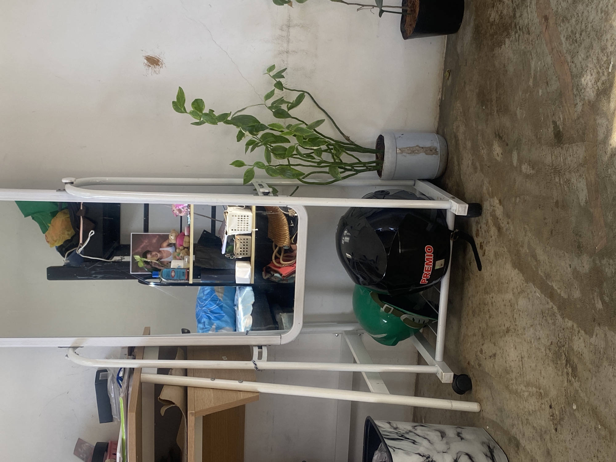 This image captures a corner of a casual indoor or semi-enclosed space, possibly in a home or utility area in Davao City, Philippines. Dominating the left side is a full-length mirror with a white metal frame. Reflected in the mirror are items on a shelf, including a framed picture of a young woman with long hair, small figurines, various baskets, and a blue bag.
To the right of the mirror, resting on its stand, are two helmets: a black one prominently displaying "PREMIO" in red text, and a green one tucked beneath it. A potted plant with green leaves in a grey and white pot stands nearby. Another plant in a dark brown pot is partially visible in the upper right. The walls are light-colored, showing some cracks and a brownish patch of discoloration. The floor is concrete, appearing worn, stained, and possibly dirty. A small wooden table or desk is partially visible in the lower left, and a black and white marbled bin is in the lower right. Bright natural light illuminates the scene, suggesting it is daytime. No specific activity is occurring.
