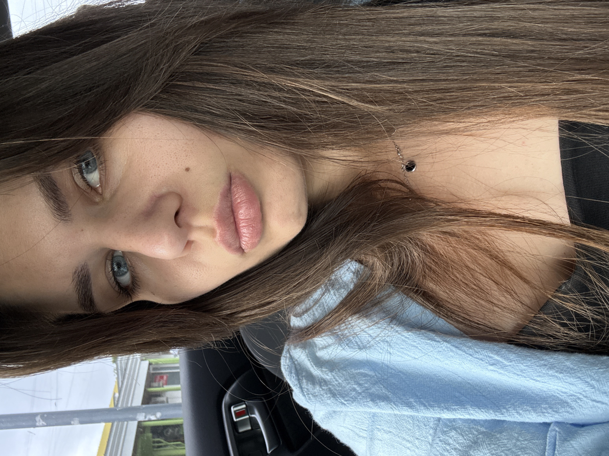 A close-up selfie shows a young woman with long, dark brown hair and striking blue eyes. She is wearing a light blue collared shirt over a black top and a delicate silver necklace with a small black pendant. The background appears to be the interior of a car, with a black door handle visible on the left side. Outside the car window, a street scene is visible with buildings and a cloudy sky. The woman's face is the primary focus, with her lips slightly parted, creating a pouty expression. The lighting is soft and natural, suggesting an indoor or overcast outdoor setting. The image captures a casual, everyday moment.