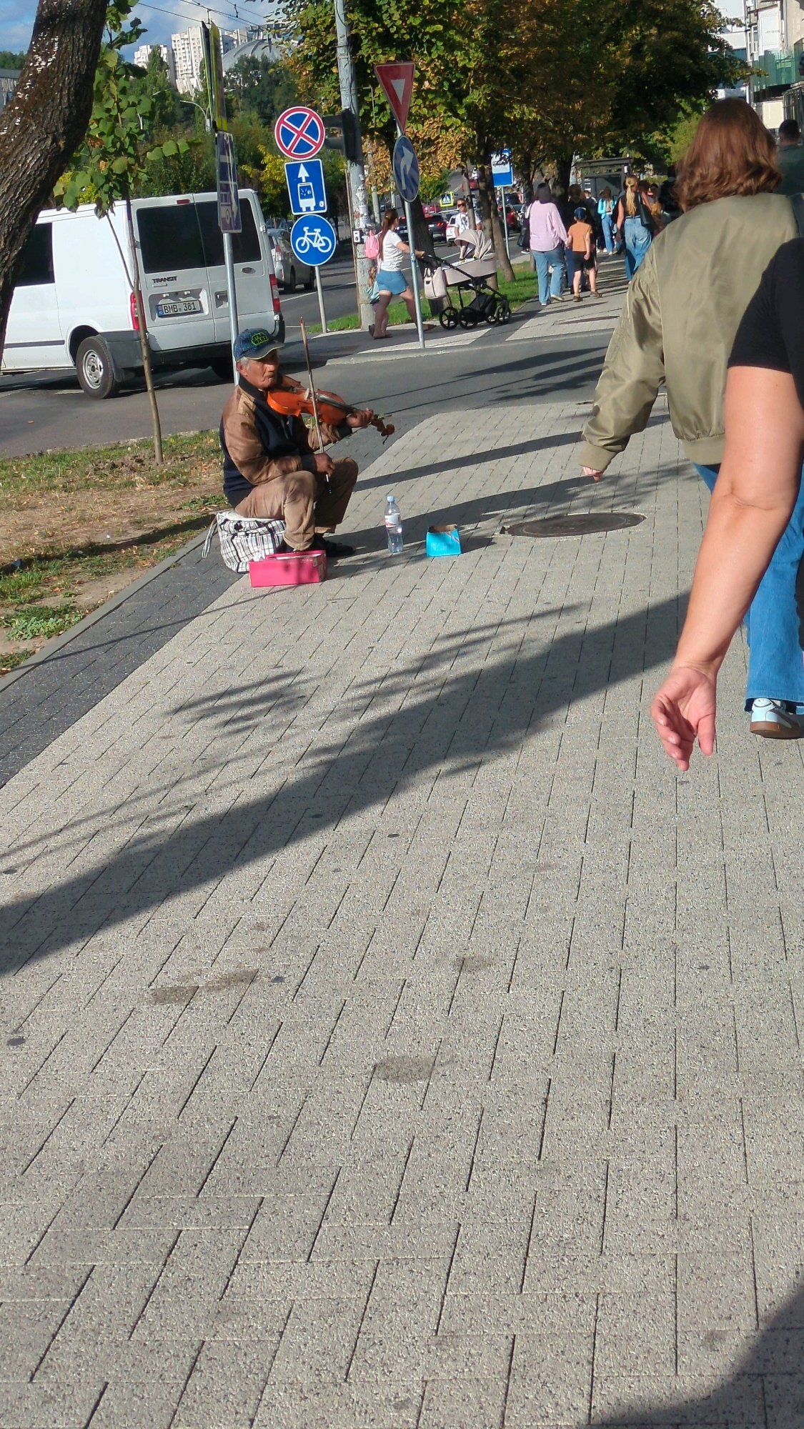 The image depicts a street scene in Chișinău, Moldova, likely in the autumn, with trees displaying fall foliage. A man is seated on a checkered cloth on the sidewalk, playing a violin. He is wearing a brown jacket and light-colored pants. In front of him, on the sidewalk, are a pink box, a bottle of water, and a blue box, suggesting he is busking for money.
Behind the violinist, a white van is parked on the street. Further back, several people are walking, including a woman pushing a stroller. Traffic signs, including one with a bicycle symbol, are visible on a pole near the sidewalk. The sidewalk is paved with gray bricks. Shadows from trees and people are cast across the ground, indicating it is daytime with a clear sky. The overall impression is a typical urban street scene with a musician performing.
