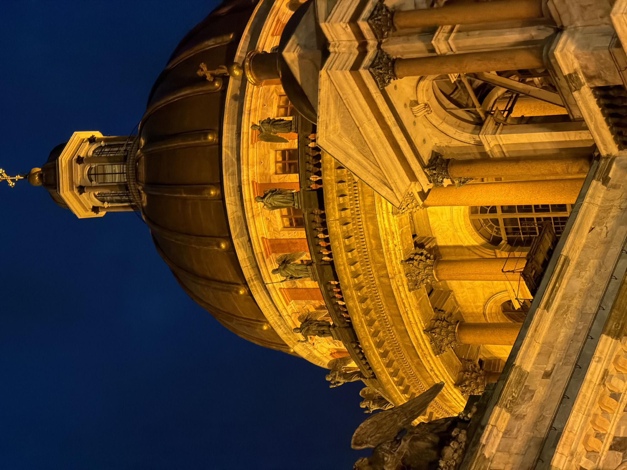 The image shows a detailed, low-angle nighttime view of the dome and upper sections of St. Isaac's Cathedral in Saint Petersburg, Russia. The ornate architecture is illuminated by artificial warm yellow light, highlighting the copper dome, the colonnaded drum with arched windows, and the statues of angels and figures adorning the upper facade. The sky is a deep, dark blue, indicating late evening or night. Architectural features such as columns with Corinthian capitals, decorative cornices, and balustrades are clearly visible. Several statues of winged figures and human-like figures are positioned along the upper parapet and on the dome's structure, some holding staffs or objects. A prominent golden cross tops the lantern at the apex of the main dome. The overall impression is one of grandeur and solemnity.