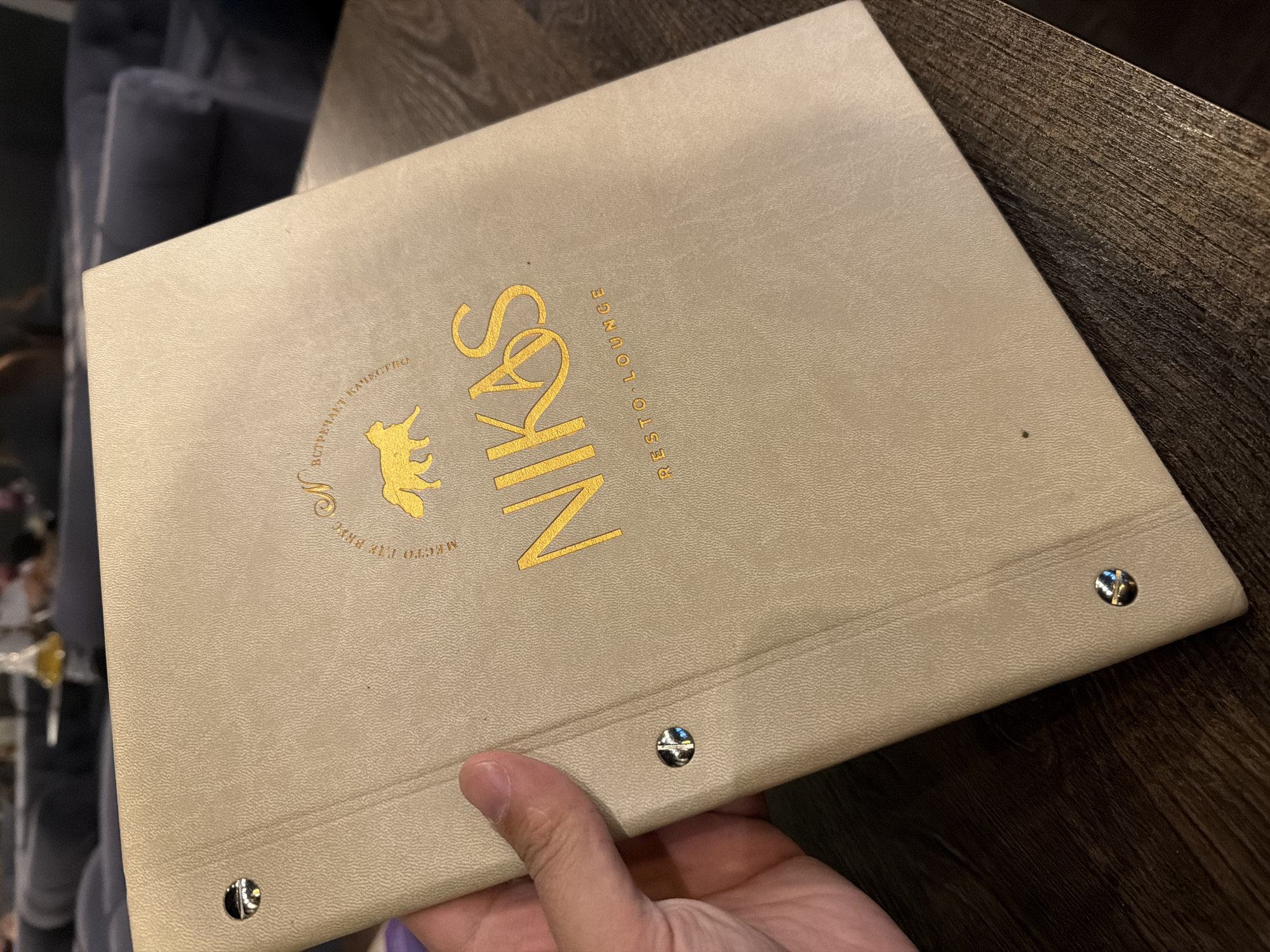 The image shows a beige menu folder from the restaurant "Nikos Resto Lounge" in Krasnoyarsk, Russia. The logo and name are printed in gold. The menu is held by a person's hand and sits on a wooden table. A blurry couch is visible in the background.