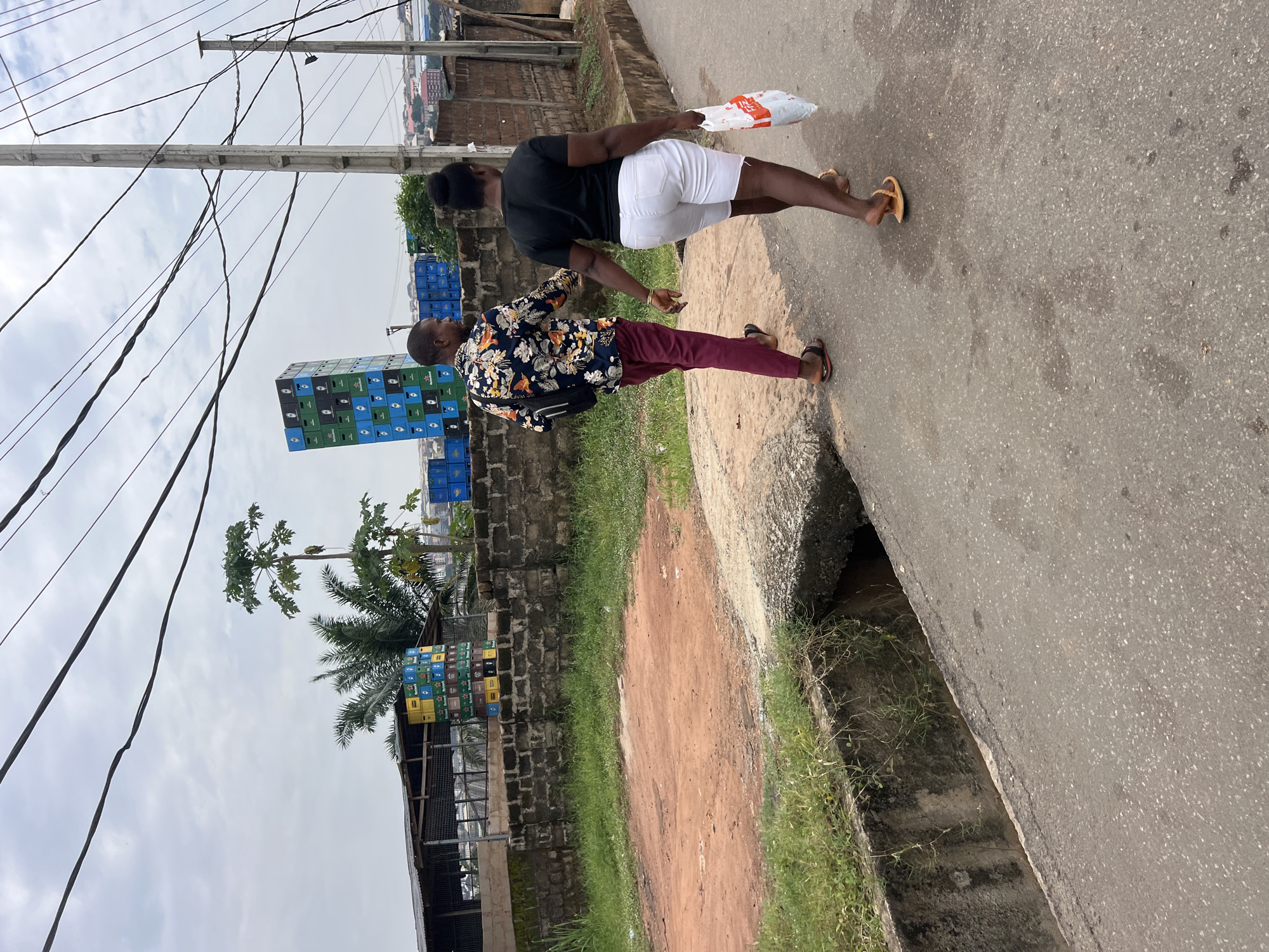 In Nkpor, Nigeria, two individuals walk away from the camera down a paved road. The person on the left is a male wearing a floral shirt and maroon pants. The person on the right is a female wearing a black t-shirt and white shorts, carrying a white plastic bag. Behind them, stacks of beer crates are piled high. The environment includes a drainage ditch on the left side of the road, a brick wall, vegetation, and utility poles with overhead wires. The sky is overcast.