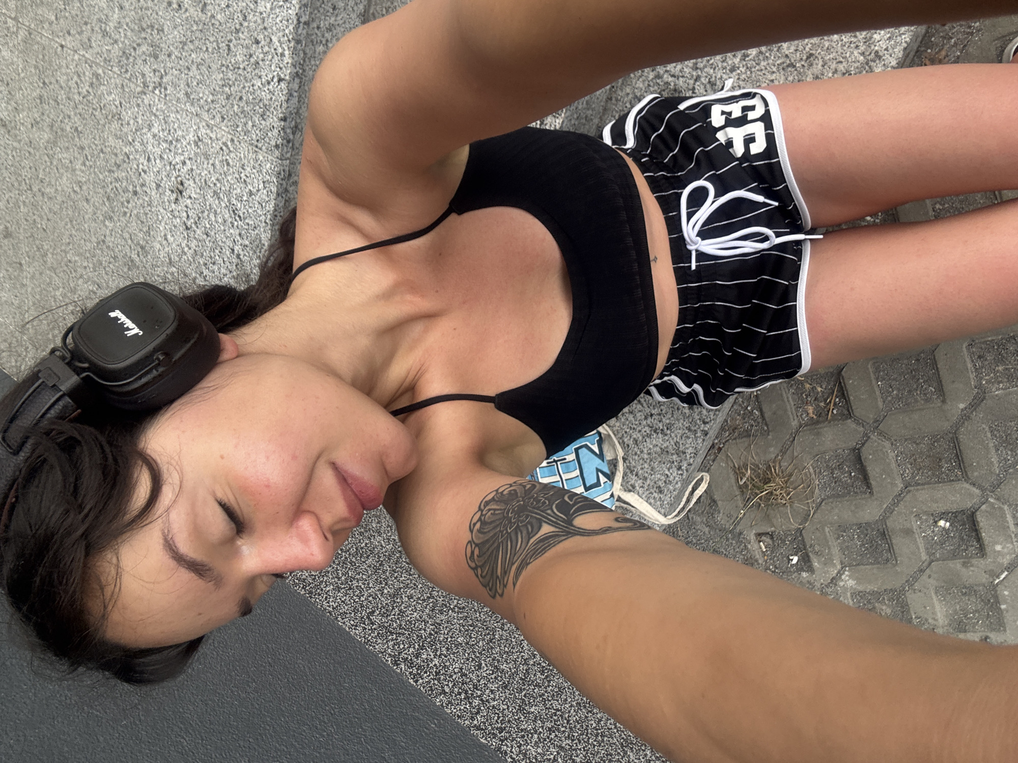 A young woman with dark hair, wearing black headphones and a black sports bra, poses for a selfie outdoors. She is dressed in black and white striped shorts with the number "33" on the right leg. She has a tattoo on her left arm. The background consists of a gray textured wall and a stone-paved ground with some dry grass. The lighting suggests it is daytime. The location is Burgas, Bulgaria.