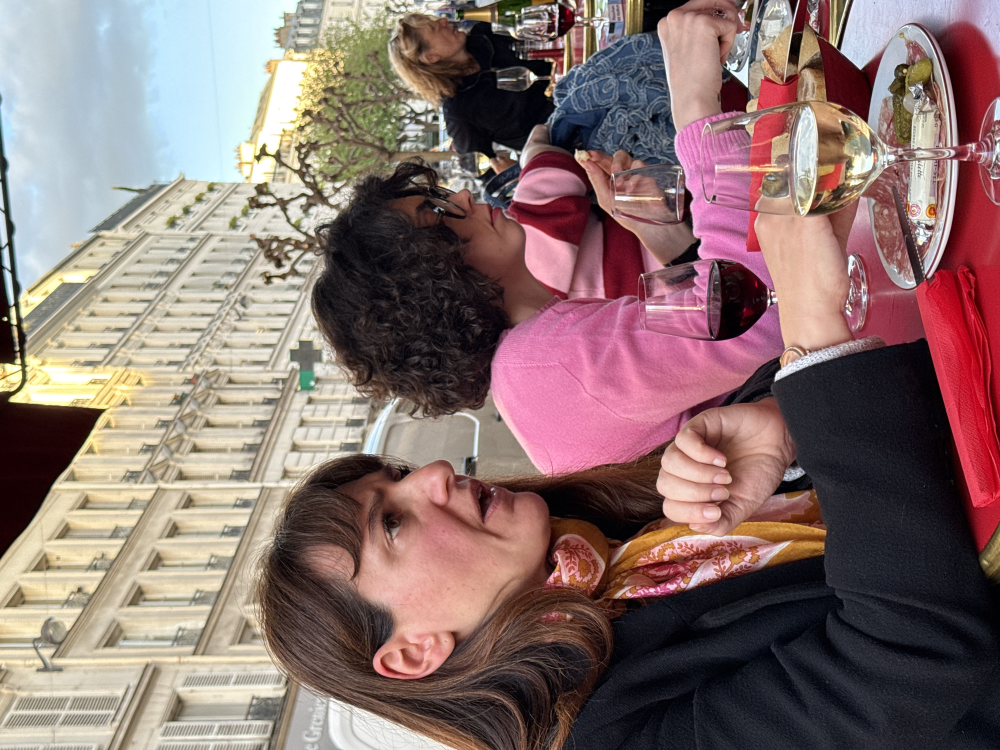 This image captures an outdoor dining scene in what appears to be Paris, France, during late afternoon or early evening. Three women are seated at a red table, enjoying drinks and charcuterie. The light is soft and warm, characteristic of the golden hour.
In the foreground, a woman with long, dark brown hair, wearing a black jacket and a patterned yellow and pink scarf, looks upwards and to the side, her mouth slightly ajar as if speaking or listening intently. Next to her, another woman with dark, curly hair and glasses is seen in profile, wearing a pink and red striped sweater under a pink long-sleeved top. She holds a glass of red wine and a piece of bread, looking towards the right. Further in the background, a third woman with blonde hair and a black top is partially visible, also seated at the table.
The table is set with glasses of red and white wine, and a champagne bottle can be seen in the background. A plate features slices of charcuterie, cornichons (pickles), and a small packet of butter, clearly labeled "BEURRE" and "BEURRETTE" with "1/2 SEL" also visible. Bread is also present on the table.
The setting reveals classic Parisian architecture, with a tall, light-colored building featuring multiple windows and balconies dominating the background. Bare tree branches suggest either early spring or late autumn. The sky is a clear light blue with some scattered clouds. The overall mood appears relaxed and social, as the women gather for a meal or drinks.