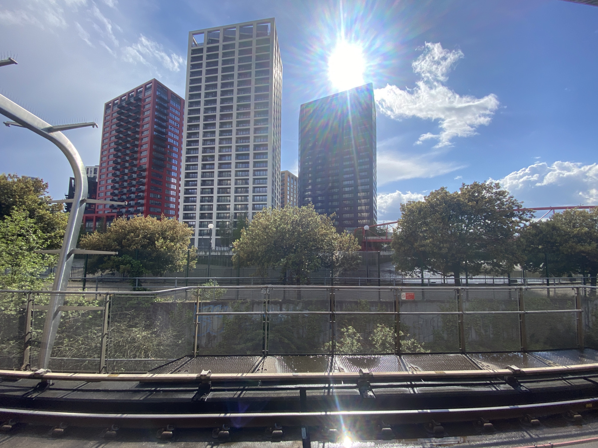 The image shows a view from a train on the Docklands Light Railway (DLR) in London, United Kingdom. Three high-rise residential buildings are visible in the background. The building on the left is red, the middle building is white with an open top, and the building to the right is a darker color and partially obscured by the sun's glare. A metal safety barrier and a line of trees separate the train tracks from the buildings. Overhead gantry and a red bridge are visible in the background to the right. The sky is blue with scattered clouds.