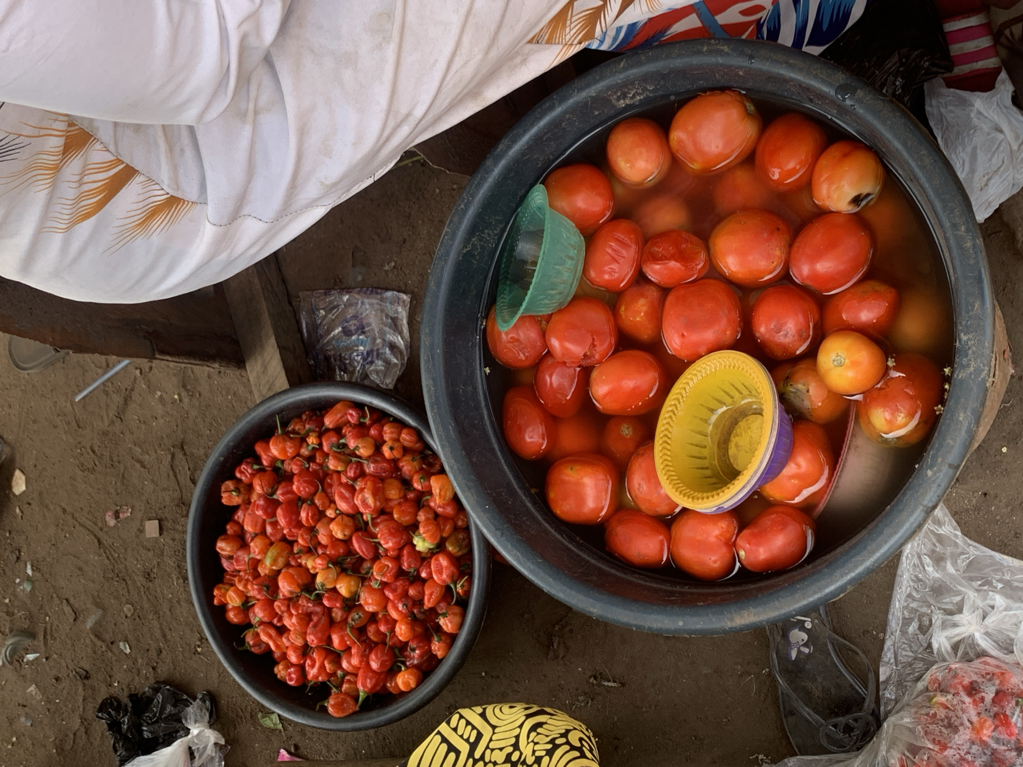 This overhead shot depicts a vibrant market scene in Ilorin, Nigeria, focusing on fresh produce laid out for sale on an earthy, dirt ground. The setting suggests an informal outdoor market, with scattered plastic bags and debris visible on the ground.
The primary subjects are two black plastic basins. The larger basin on the right contains numerous red tomatoes submerged in water. Many of the tomatoes appear ripe, with some showing variations in color from deep red to orange, and a few exhibiting slight imperfections or softened spots. Two small, colored plastic scoops, one green and one yellow with a purple rim, float among the tomatoes, likely for serving or measuring. The practice of keeping tomatoes in water is common in warm climates to maintain freshness and cool the produce.
To the left, a smaller black basin is brimming with a large quantity of small, intensely red and orange peppers, resembling Scotch bonnets or similar hot pepper varieties popular in the region.
The bottom right corner of the image shows a partial view of a person's foot, clad in a dark flip-flop or sandal, indicating human presence, likely a vendor or customer. A piece of yellow fabric with a black, geometric pattern is visible at the very bottom center, possibly part of clothing or a bag. Along the top edge, a white fabric with an orange and dark blue pattern suggests either part of a stall covering, an awning, or another person's attire.
The activity taking place is a market transaction, showcasing fresh vegetables available for purchase. The lighting suggests it is daytime, though no specific time of day or weather conditions can be definitively determined. There is no visible text in the image.