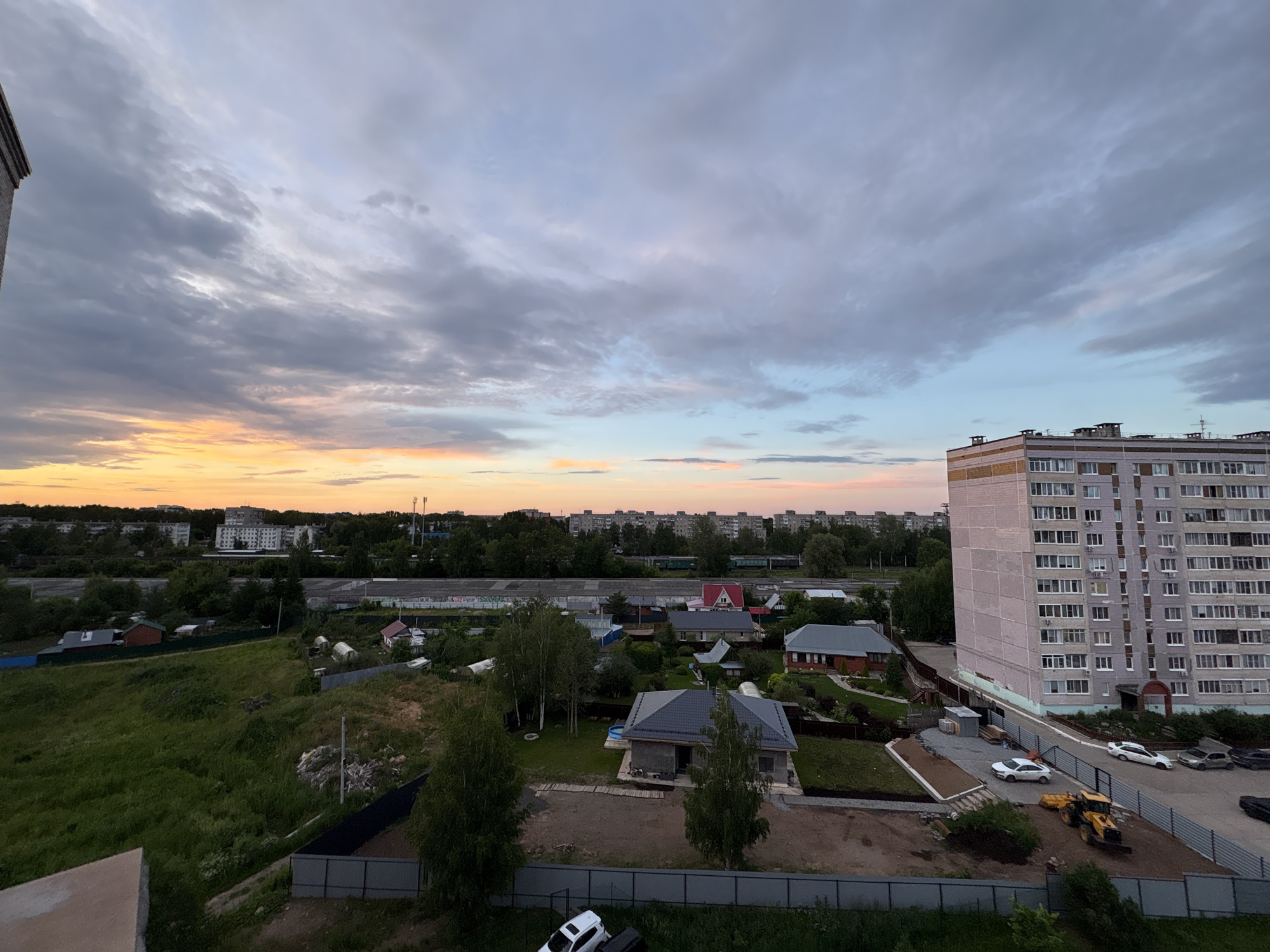 This is an aerial, wide-angle photograph of a suburban area in Chaikovsky, Russia, at dusk. The sky is filled with clouds, with hues of orange and pink near the horizon transitioning to light blue and gray overhead. In the foreground, to the left, a portion of a residential building's facade is visible.
To the right, a tall, multi-story apartment building with a light pinkish-beige facade dominates the scene. It has numerous windows and visible chimneys on its roof. A fence runs along the base of the apartment building and extends into the foreground. Several cars are parked near the building, including a white sedan, another white vehicle, and a dark-colored SUV. A yellow backhoe loader is also present on the construction site.
Further into the middle ground, a collection of smaller residential buildings and houses with fenced yards are scattered across a grassy, uneven terrain. Some of these houses appear to be under construction, with exposed foundations and building materials. Trees and various types of vegetation are interspersed throughout the area. A railway track with green train cars is visible in the mid-distance, separating the residential area from a line of larger, apartment-style buildings in the background. The overall impression is a developing residential area with a mix of older apartment blocks and newer, single-family homes.