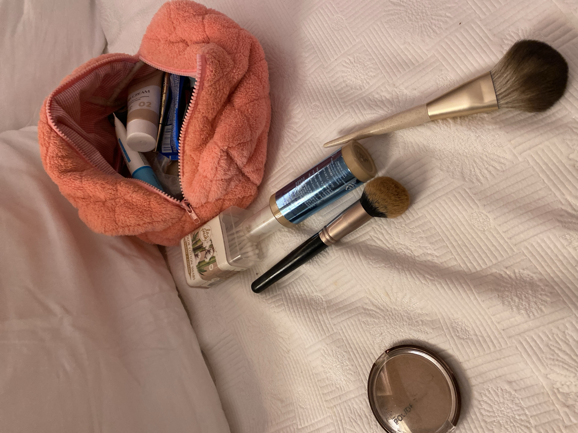 A peach-colored fuzzy makeup bag is open on a white, textured surface, spilling out various makeup items. Visible items include a bottle of foundation, two makeup brushes, a compact of powder, and a box of cotton swabs. The scene is illuminated by soft, ambient light. No people are present, and the background consists of a blurred white surface, possibly a bed. The image does not provide any information or visual cues to suggest its location in Aksu, Turkey.