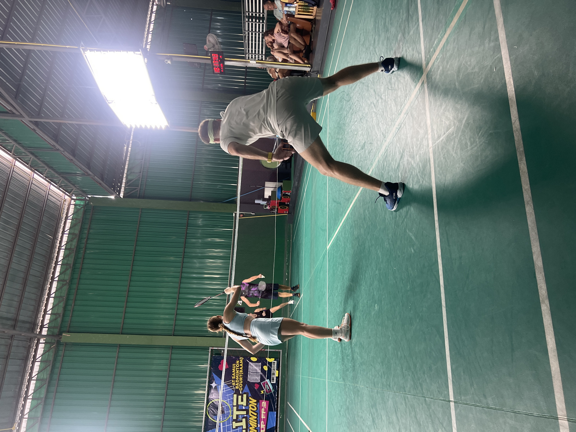 An indoor badminton court is depicted with four people playing a game. In the foreground, a man in a light-colored t-shirt and grey shorts is positioned in a ready stance on the right side of the frame, facing away from the camera. On the left side of the foreground, a woman in a light blue sports bra and skirt is also facing away from the camera, holding a badminton racket. In the middle ground, a man in a dark purple and black patterned t-shirt and dark shorts is visible, and further back, another player in a dark outfit can be seen. The court is green with white lines marking the boundaries. The background shows a green metal wall and a brightly lit area with a ceiling structure. Spectators are seated in the background on the right side, observing the game. A scoreboard with red digital numbers is mounted on a pole to the right.