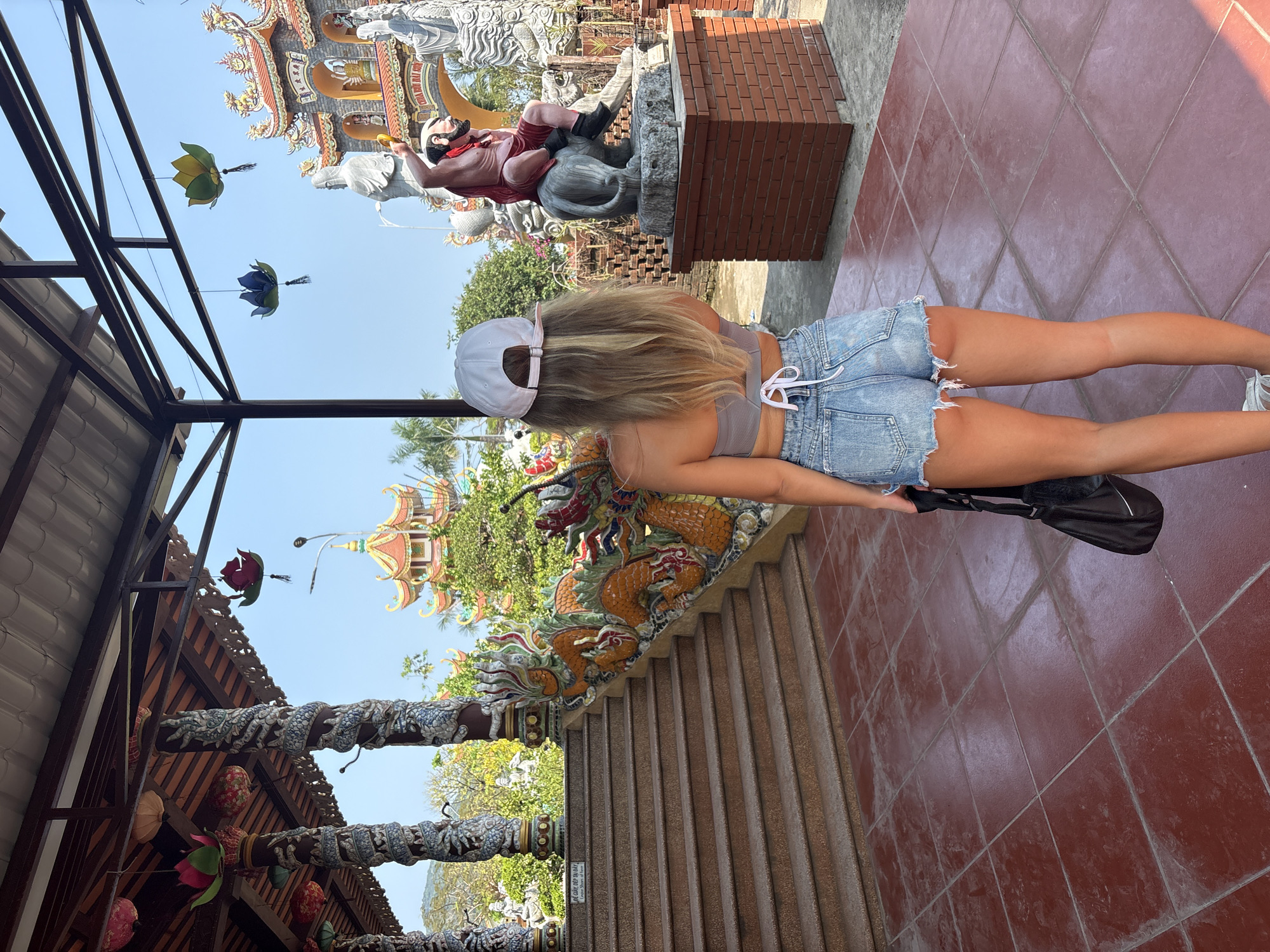 A bright daylight scene captures a young woman from behind, exploring an ornate temple or pagoda complex in Đà Nẵng, Vietnam. She has long blonde hair and wears a white baseball cap, a grey crop top, and light blue denim cut-off shorts, carrying a black bag in her left hand. She is walking on a red-tiled path towards what appears to be a set of stairs.
The setting is rich with cultural and artistic details. To the woman's right, a large statue features a figure in red and white attire, possibly a deity or historical figure, riding an animal that resembles an ox or buffalo, placed on a grey stone base next to a brick structure. Further in the background, more large, lighter-colored statues and a multi-tiered, intricately decorated temple gateway with ornate roofing are visible under a clear blue sky.
To her left, a balustrade or low wall is adorned with vibrant, relief sculptures of dragons in hues of orange, green, and blue. Overhead, several lotus-shaped lanterns in green, blue, and red hang from dark metal beams, adding to the decorative ambiance. Beyond these structures, palm trees and other greenery suggest an open-air environment. The overall impression is one of a sunny, pleasant day, ideal for sightseeing and cultural immersion. Some indistinct text is visible on a small plaque or sign near the ground, but it is too blurry to read clearly.
