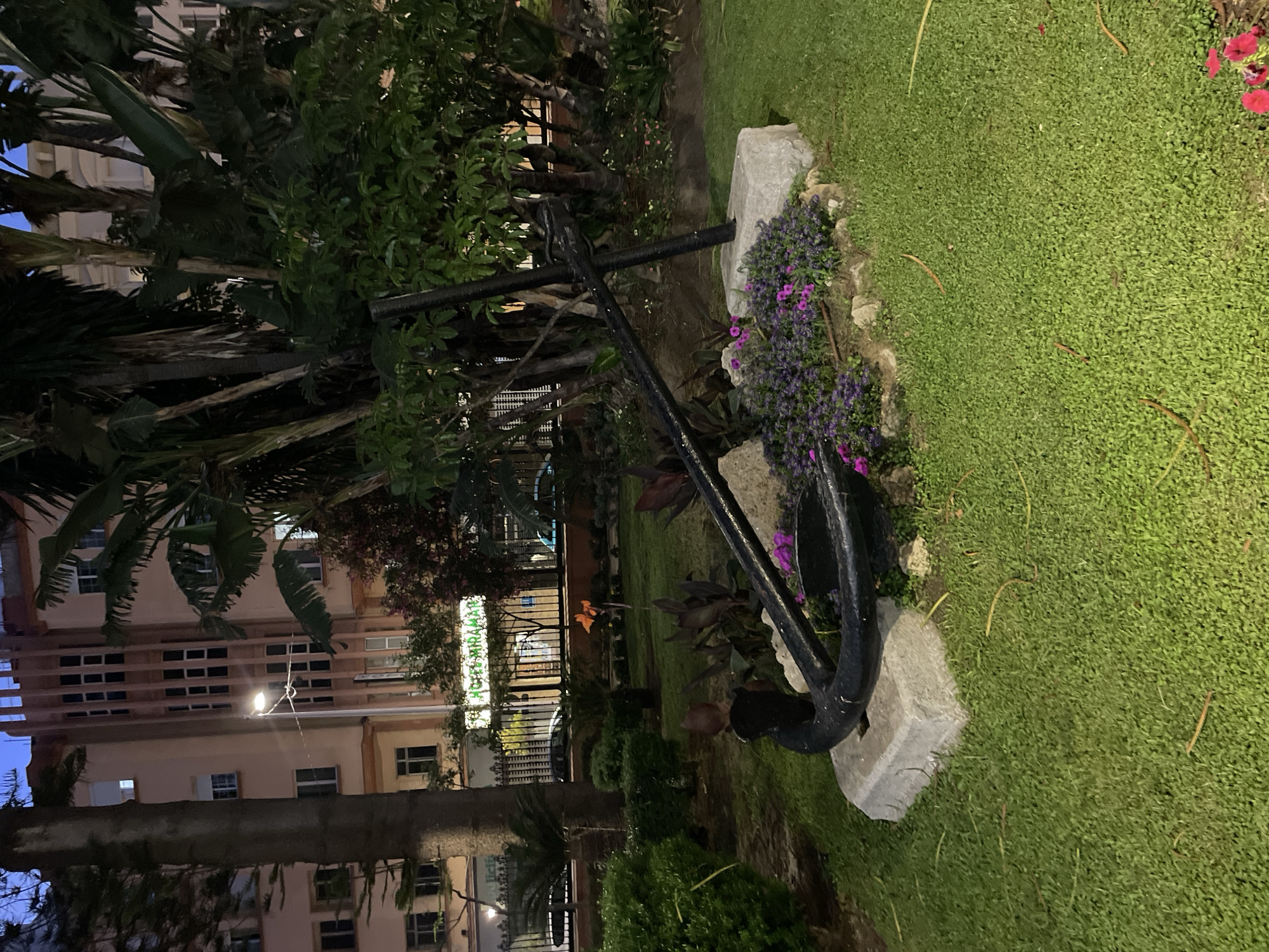 An outdoor nighttime photograph taken in La Línea de la Concepción, Spain, features an anchor as a prominent decorative element in a garden setting. The anchor, made of black metal, rests on a concrete base and is partially surrounded by small purple flowers. The garden is neatly mowed, with green grass covering the ground. Behind the garden, a multi-story building with a distinctive Art Deco architectural style is visible. The building's facade is painted in a light peachy-pink hue, with numerous rectangular windows. A street lamp illuminates the scene, casting a warm glow. In the background, a fence separates the garden area from the street, beyond which cars are parked and the entrance to "Hotel Miramar" is clearly lit and visible. Tropical-looking plants, including palm trees and large-leafed foliage, are interspersed throughout the garden, adding to the lush environment. The sky above is a deep twilight blue, indicating the late hour.