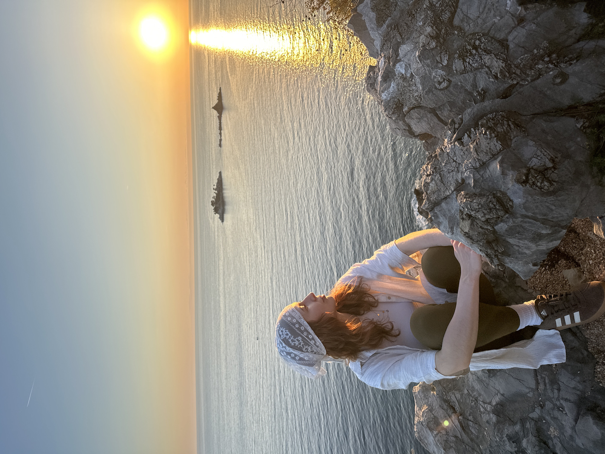 A woman sits on a rocky cliff overlooking the sea at sunset in Kaluđerac, Montenegro. The sun is visible on the horizon, with a bright reflection on the water. Two small rocky islands are visible in the distance. The woman is wearing a white shirt, green pants, and a white headscarf. She is looking out at the sea. She is wearing brown adidas shoes with white stripes.