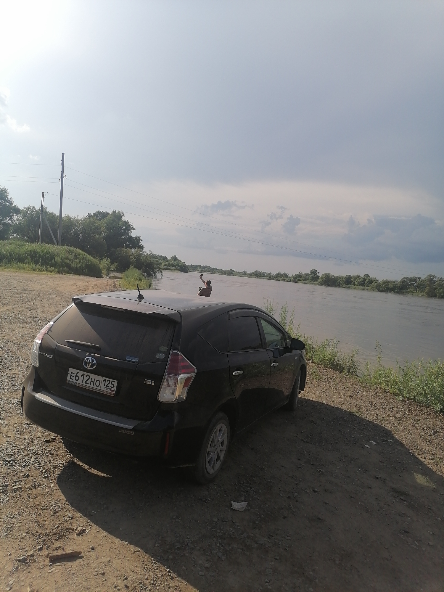 A black Toyota Prius is parked on a dirt road next to a wide, calm river under a cloudy sky. A person is standing in the shallow water near the riverbank, facing away from the camera and holding a smartphone up, as if taking a photo or video. Green trees and vegetation line the far bank of the river. Power lines and poles are visible in the background. The license plate of the car reads E612HO 125. The scene appears to be in a rural area, possibly on the outskirts of Novopokrovka, Russia, given the context of the location provided.