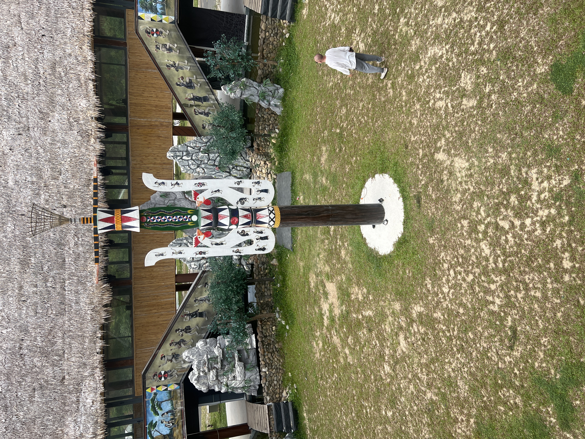The image depicts an outdoor cultural display, likely within a park or attraction in Đà Nẵng, Vietnam. In the foreground, a man wearing a white shirt and grey pants stands looking upwards. Dominating the center of the scene is a tall, intricately decorated wooden pole, adorned with colorful geometric patterns and stylized figures. This pole stands on a circular concrete base. To the left and right of the pole, tiered structures with stairs are visible, decorated with murals and sculptures depicting people in traditional attire. The background features a large building with a thatched roof, its facade partially covered by bamboo. The ground is a mixture of dry soil and sparse green grass. The overall impression is one of a cultural heritage site or theme park.