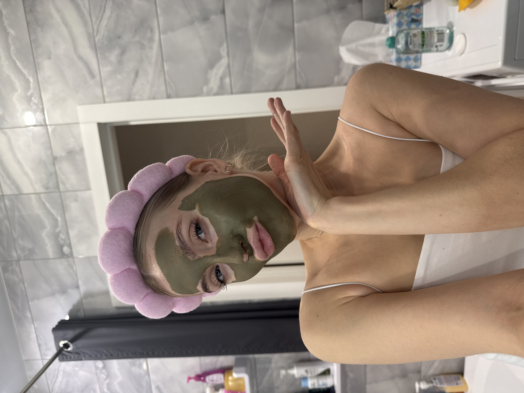 The image depicts a woman in a bathroom. She is wearing a pink headband and has a green facial mask applied. Her arms are raised, with one hand near her face. The background includes bathroom tiles, a shower curtain, and toiletries on a shelf. There is no indication of the location being Izhevsk, Russia.