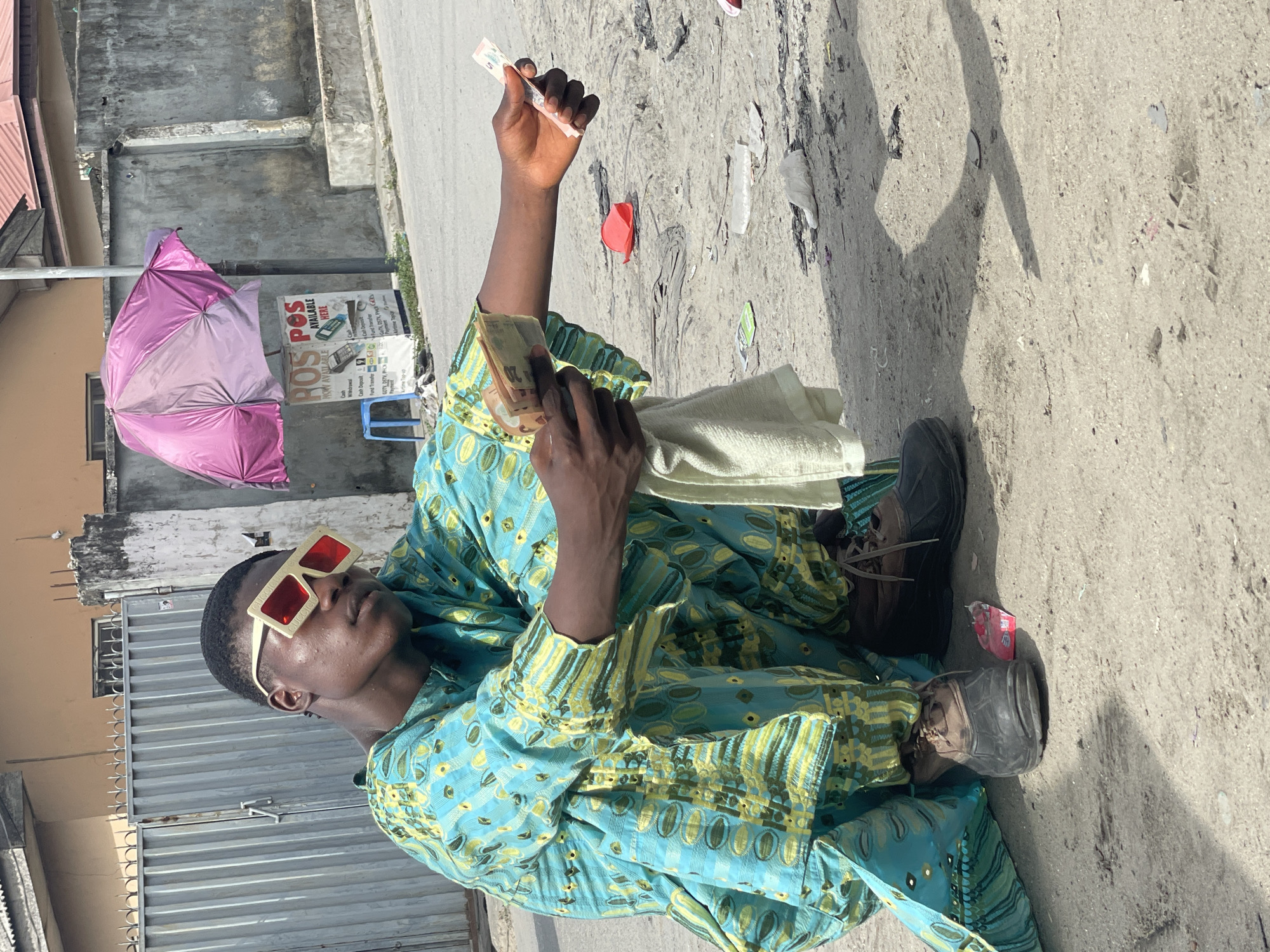 An individual is sitting on the side of a paved road in Ado, Nigeria. The person is wearing a blue patterned robe, and sunglasses, and is holding money and a cloth in their hands. The background features a building, a purple umbrella, a POS sign, and litter on the ground. The person is casting a shadow on the ground.