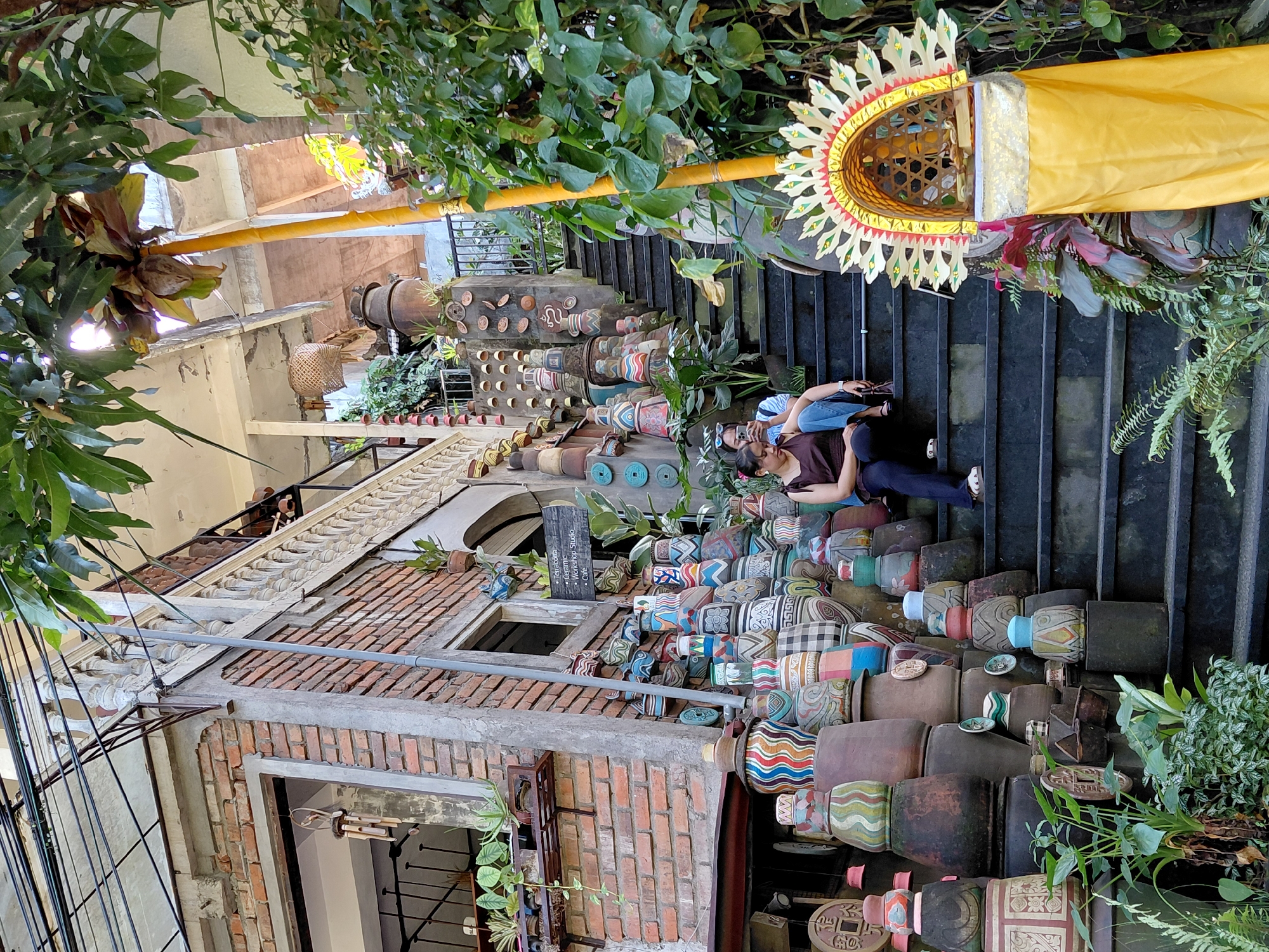 This image depicts a wide outdoor staircase in the city of Ubud, Indonesia. The staircase is flanked by numerous decorative ceramic pots of varying sizes, shapes, and intricate patterns, some stacked high. Two women are seated on the steps, facing forward. One woman, wearing a dark brown top and dark jeans, is looking down at her phone. The other woman, wearing a blue shirt and jeans, is looking away from the camera. To the right of the stairs, a traditional Balinese offering shrine, adorned with a yellow cloth and a golden, fan-like headdress, is visible. Lush green foliage surrounds the scene, with some plants growing on the stairs and along the building's exterior. The building itself appears to be made of brick, with an ornate white railing visible on an upper level. A sign on the wall reads "Terracotta Ceramic Workshop Studio Cafe".