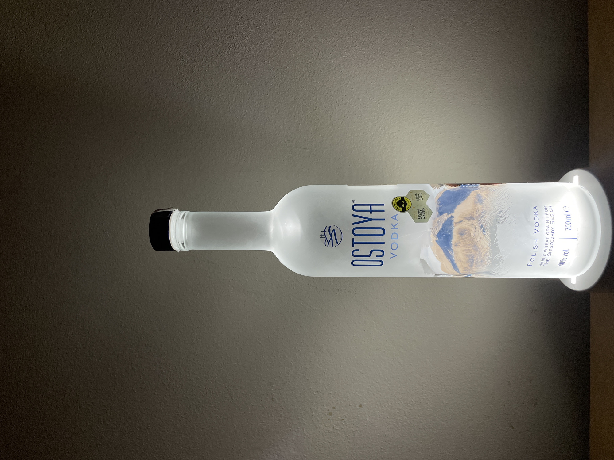 The image shows a bottle of Ostoya Vodka resting horizontally on a surface. The bottle is frosted, with a black cap and a white label. The label features the brand name "OSTOYA VODKA" in bold blue letters. The label also includes Polish Vodka details, such as "Noble wheat grain from the Bieszczady region". The alcohol content is labeled as "40% vol." The bottle is set against a plain, neutral-colored background, and the lighting is bright. There is no indication of this image being located in Sofia, Bulgaria.