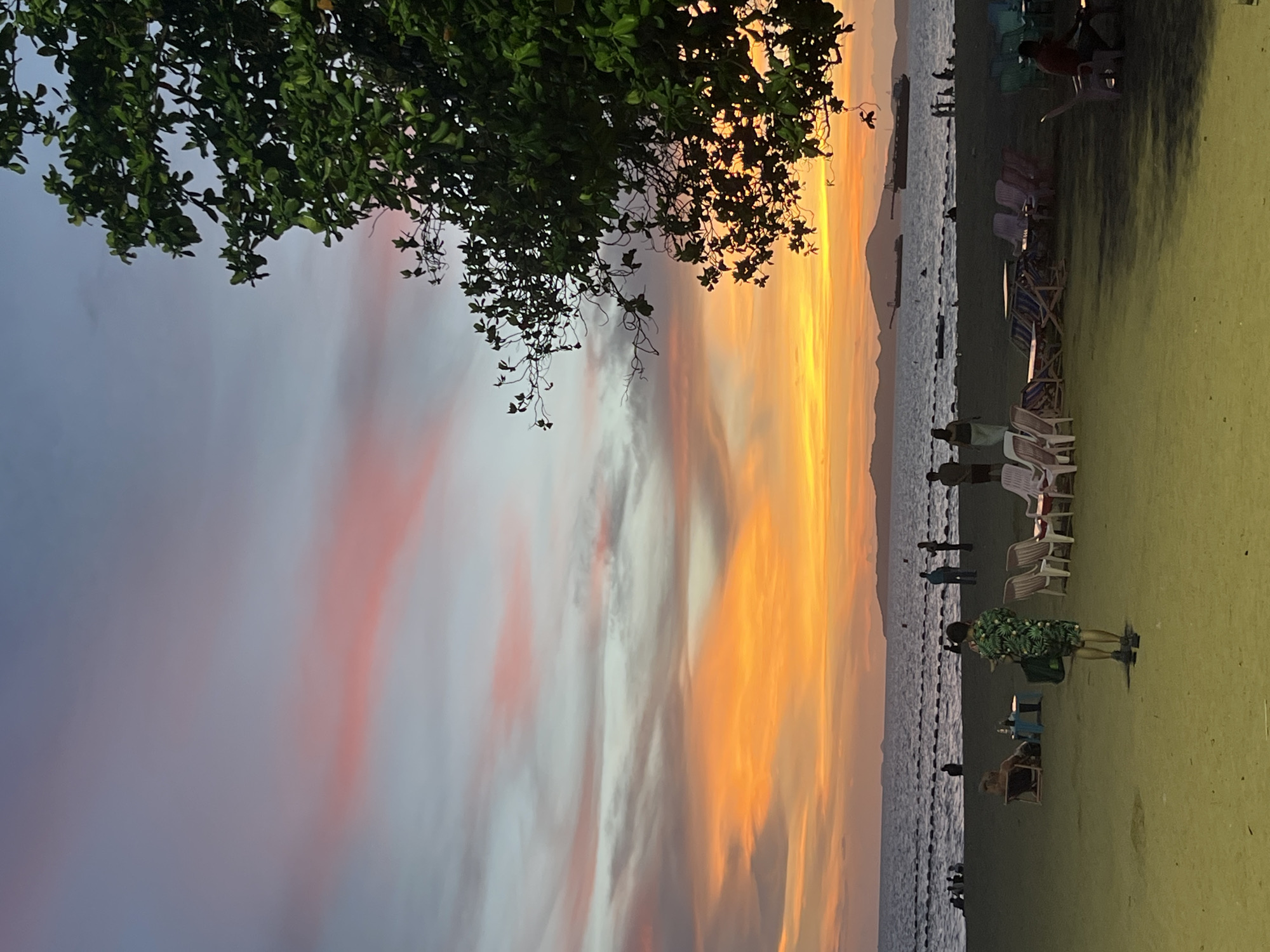 Captured in Pattaya City, Thailand, the scene unfolds at dusk. The sky is painted with hues of orange, pink, and grey clouds, with a tree branch with green leaves in the upper left of the frame. The beach transitions to a line of seated beach chairs. People meander along the shoreline and on the grass. A person wearing a shirt and shorts covered in large green leaves, faces toward the ocean. Ships sit in the water. A mountain lies to the right in the distance.