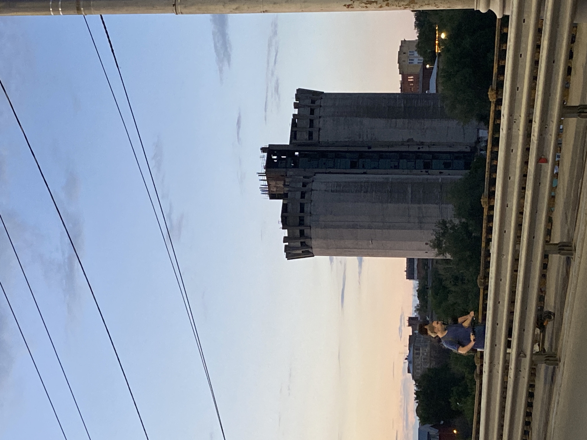 The image is a vertical shot of an industrial area at dusk. In the foreground, a metal structure with corrugated panels runs along the right side of the frame. Several parallel wires are strung across the left side of the image against a pale blue and orange sky.
Dominating the middle ground is a large, unfinished concrete building, possibly a silo or industrial tower, with visible structural elements and exposed dark openings. Trees and other buildings are visible in the background, suggesting an urban or semi-urban industrial setting.
On the right side of the frame, two people are standing near the edge of an elevated structure, possibly a bridge or walkway. One person, wearing a blue shirt, is looking towards the other person, who is partially obscured. The time of day is clearly dusk, with the sky displaying gradients of blue and soft orange hues. The overall mood is somewhat subdued, reflecting the quietude of the evening in an industrial environment. There is no visible text in the image.