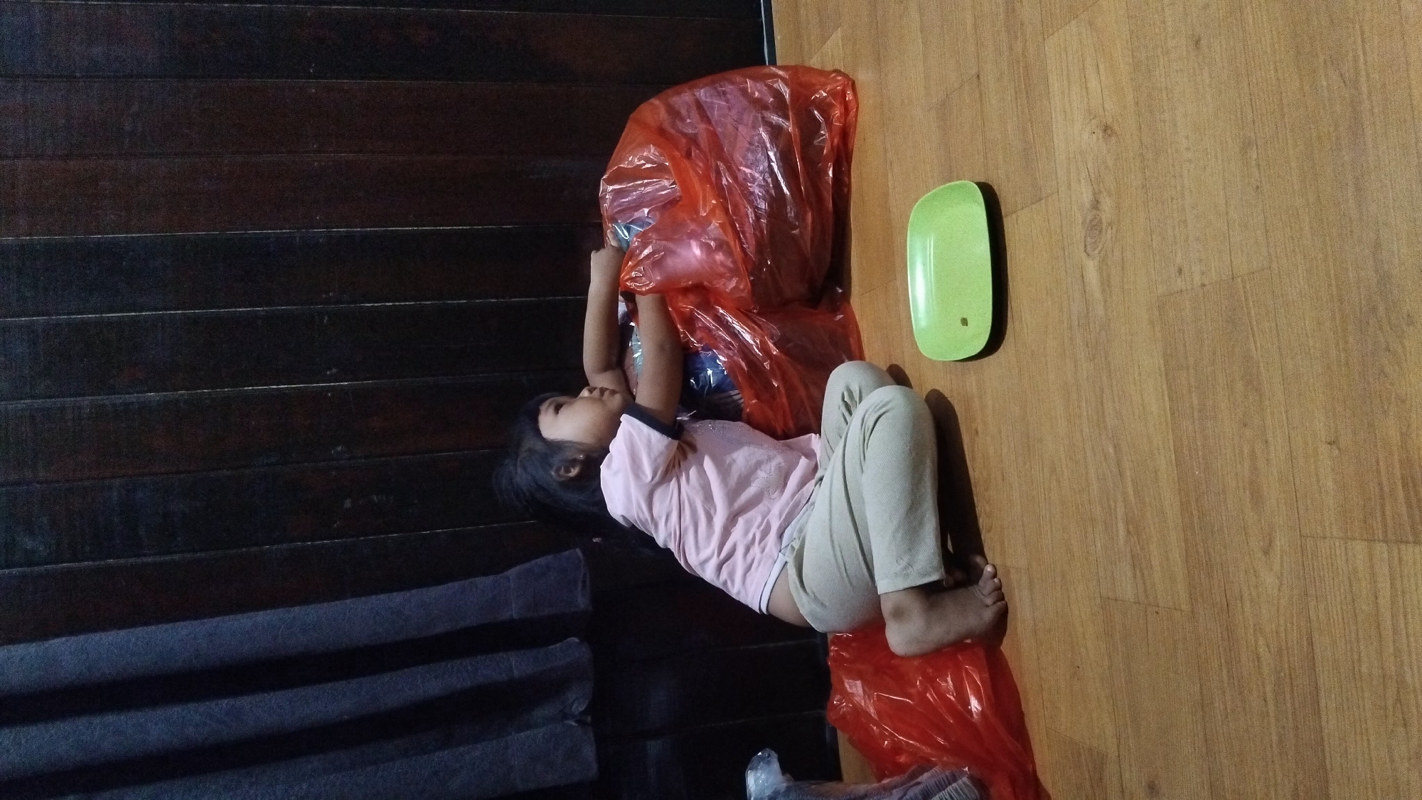 A young girl kneels on a wooden floor, her back to the viewer, looking towards a large orange plastic bag. She is wearing a pink t-shirt and beige pants. The floor has a visible wood grain pattern. To the right of the girl, a small, light green rectangular plate with rounded corners sits on the floor. In the background, a dark wooden wall with vertical planks is visible, and to the left, a dark blue curtain hangs. The scene appears to be indoors, possibly in a home in Bangun Rejo, Indonesia.