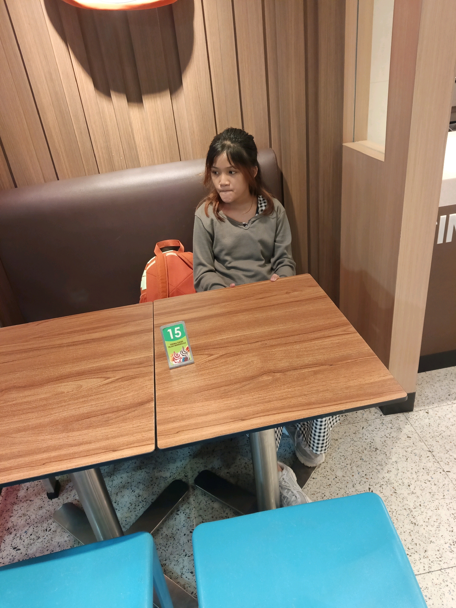 A young girl with dark hair and reddish highlights, wearing a gray long-sleeved shirt and black and white checkered pants, sits at a wooden table in a restaurant in Central Jakarta, Indonesia. An orange backpack is placed on the seat behind her. A green sign with the number "15" is on the table in front of her, along with a small promotional card featuring smiling characters. The table has a metal base and is positioned in front of a brown leather banquette. The environment includes wood-paneled walls and a speckled floor. Two bright blue chairs are visible in the foreground, partially obscuring the table. The lighting suggests an indoor setting.