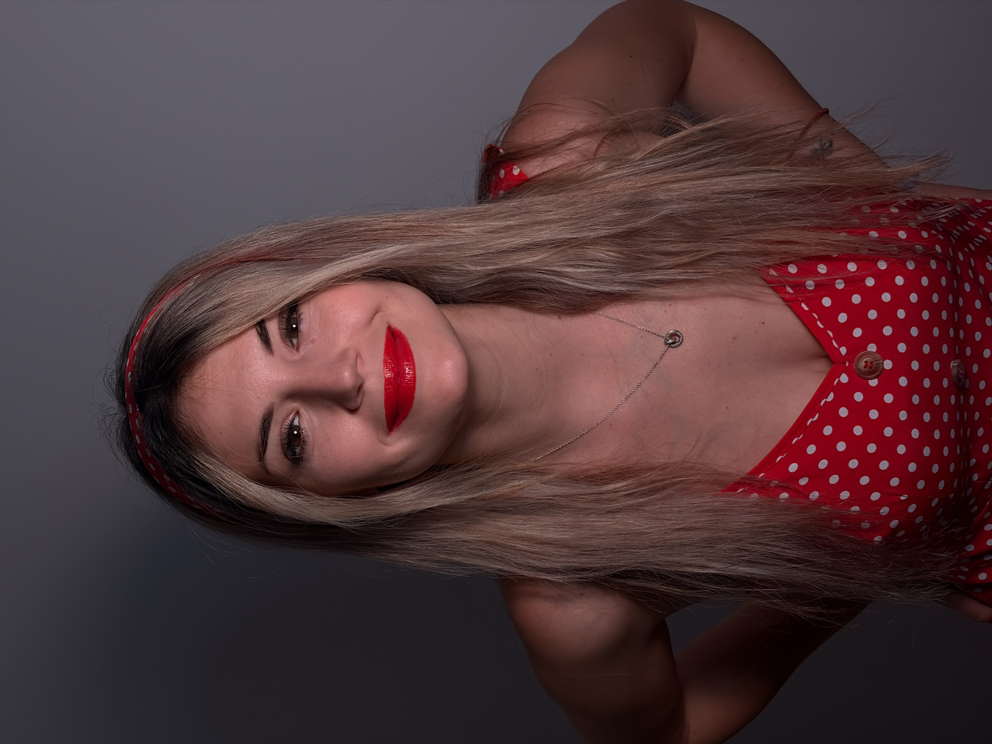 The image shows a woman with light blonde hair, wearing a red polka-dot dress and a matching headband. She is wearing red lipstick and a silver necklace. The background is a dark gray color. The image appears to be taken in a studio setting due to the plain background and controlled lighting. The location of Budva, Montenegro is not evident from the image.